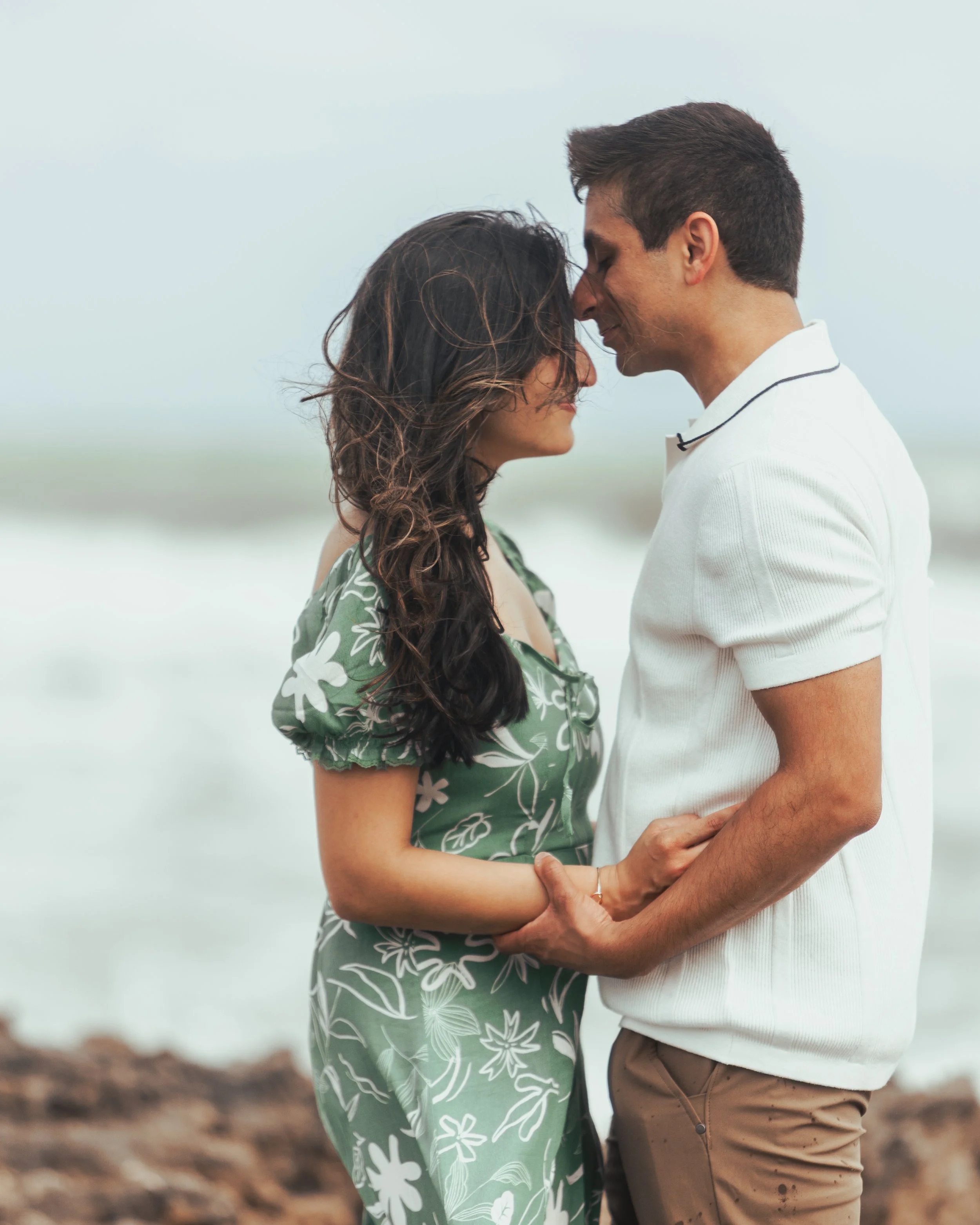 A couple embraces on a beach at sunset, with their foreheads touching and holding hands.