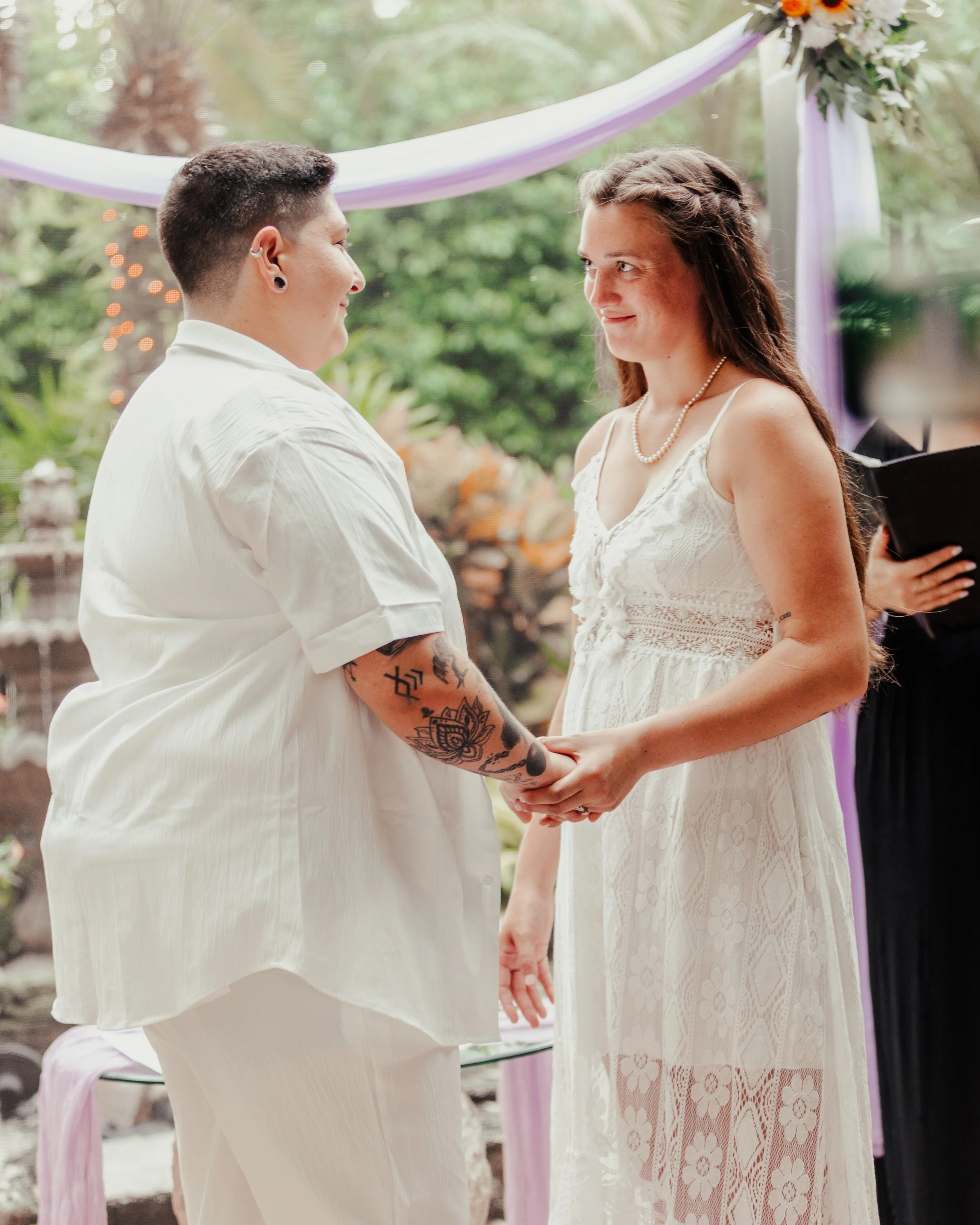Two women holding hands during a wedding ceremony outdoors, with green trees and purple and white decorations in the background.