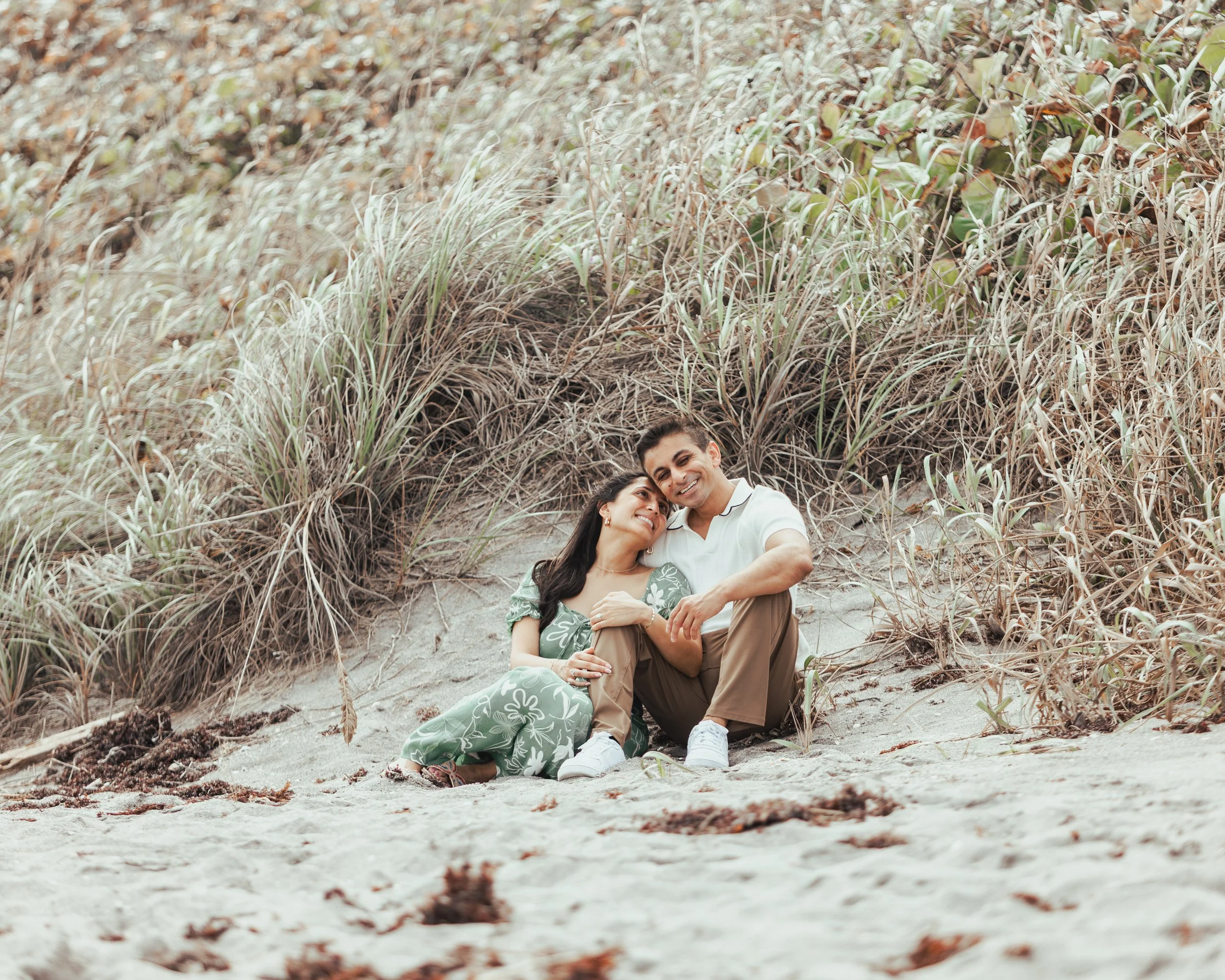 A couple sitting on the sand at the beach, smiling and cuddling with sand dunes and grass in the background.