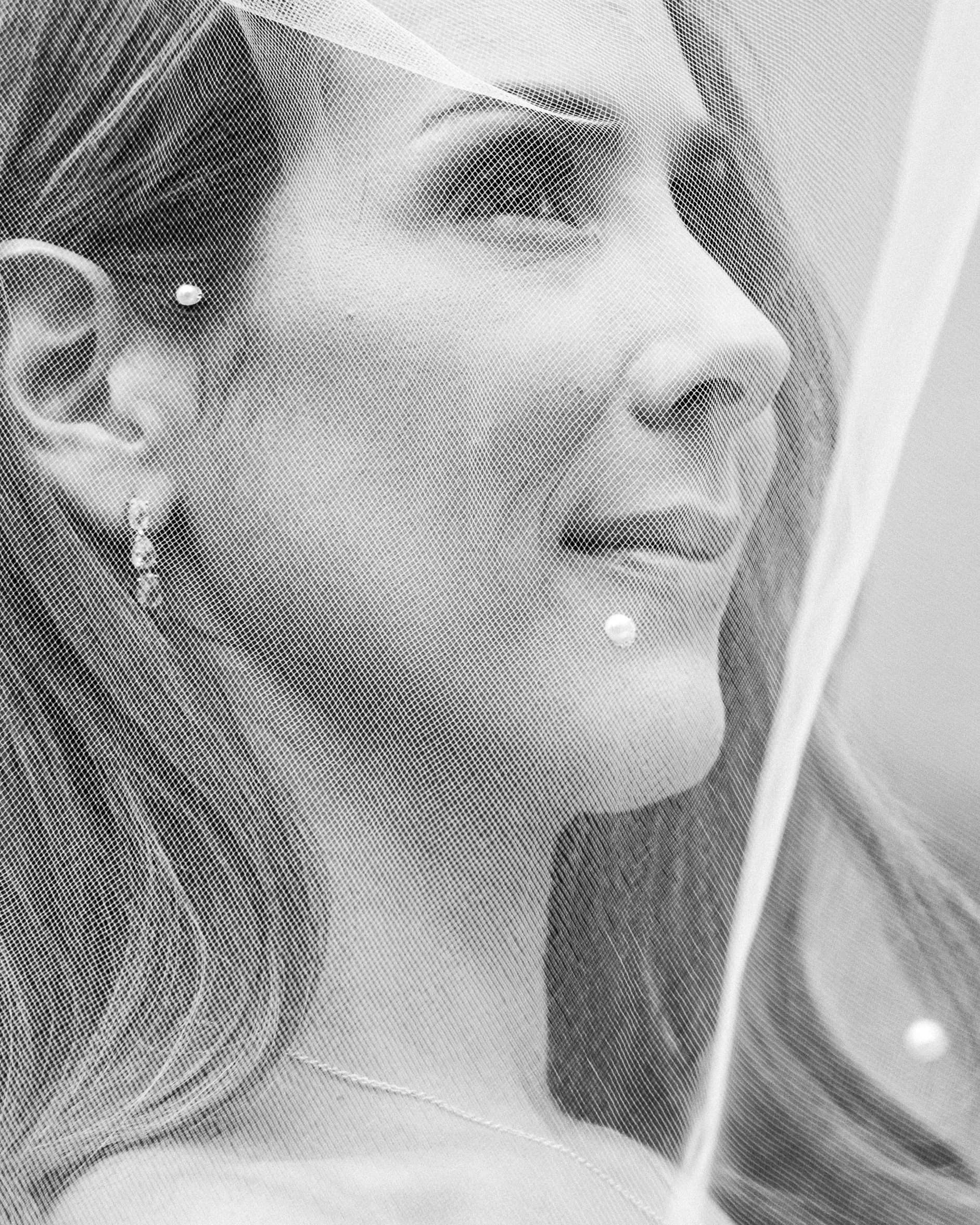 Close-up of a bride's face behind a veil, showing earrings and pearl detail in black and white.