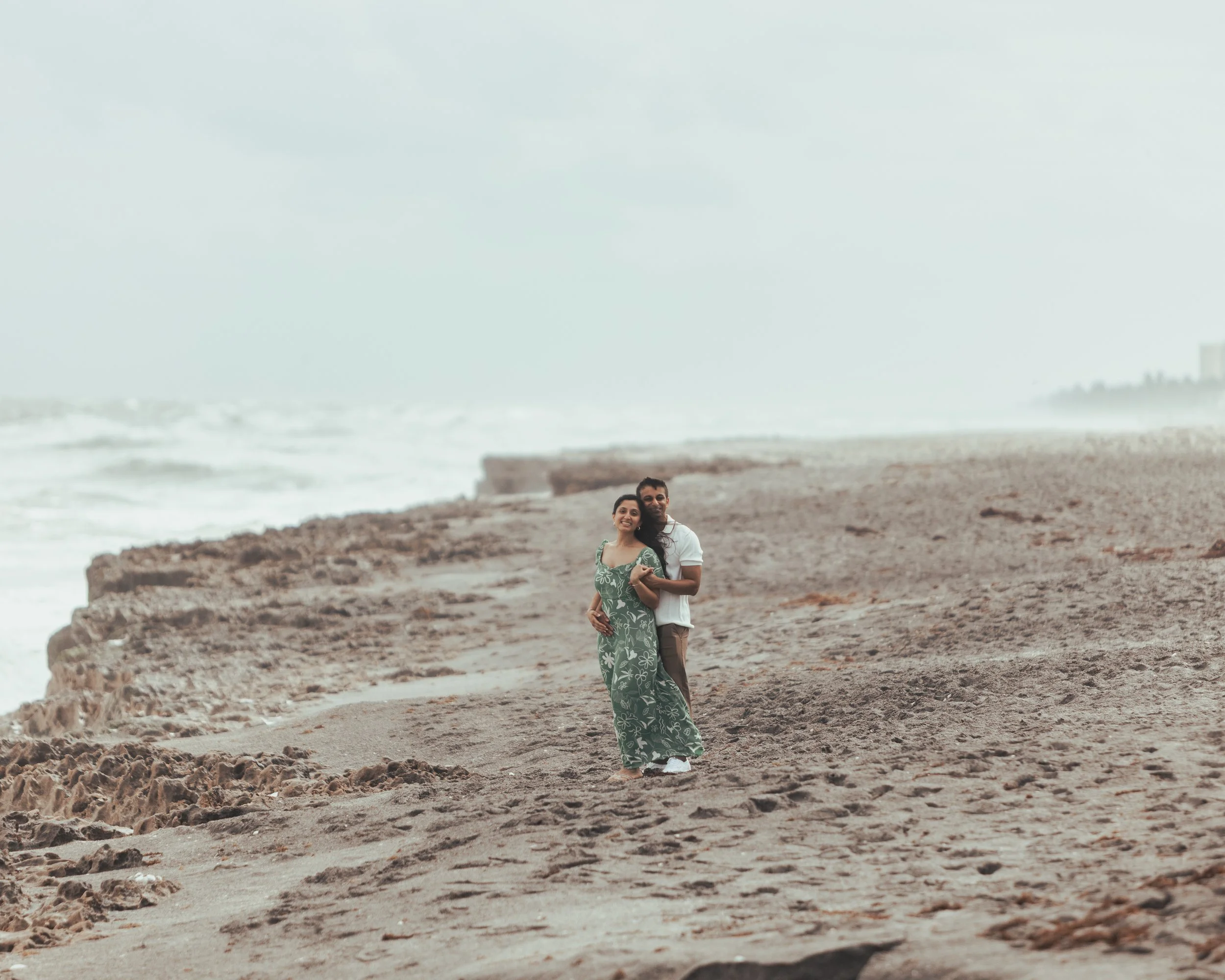A couple standing on a sandy beach, with the ocean waves in the background, smiling and embracing each other.