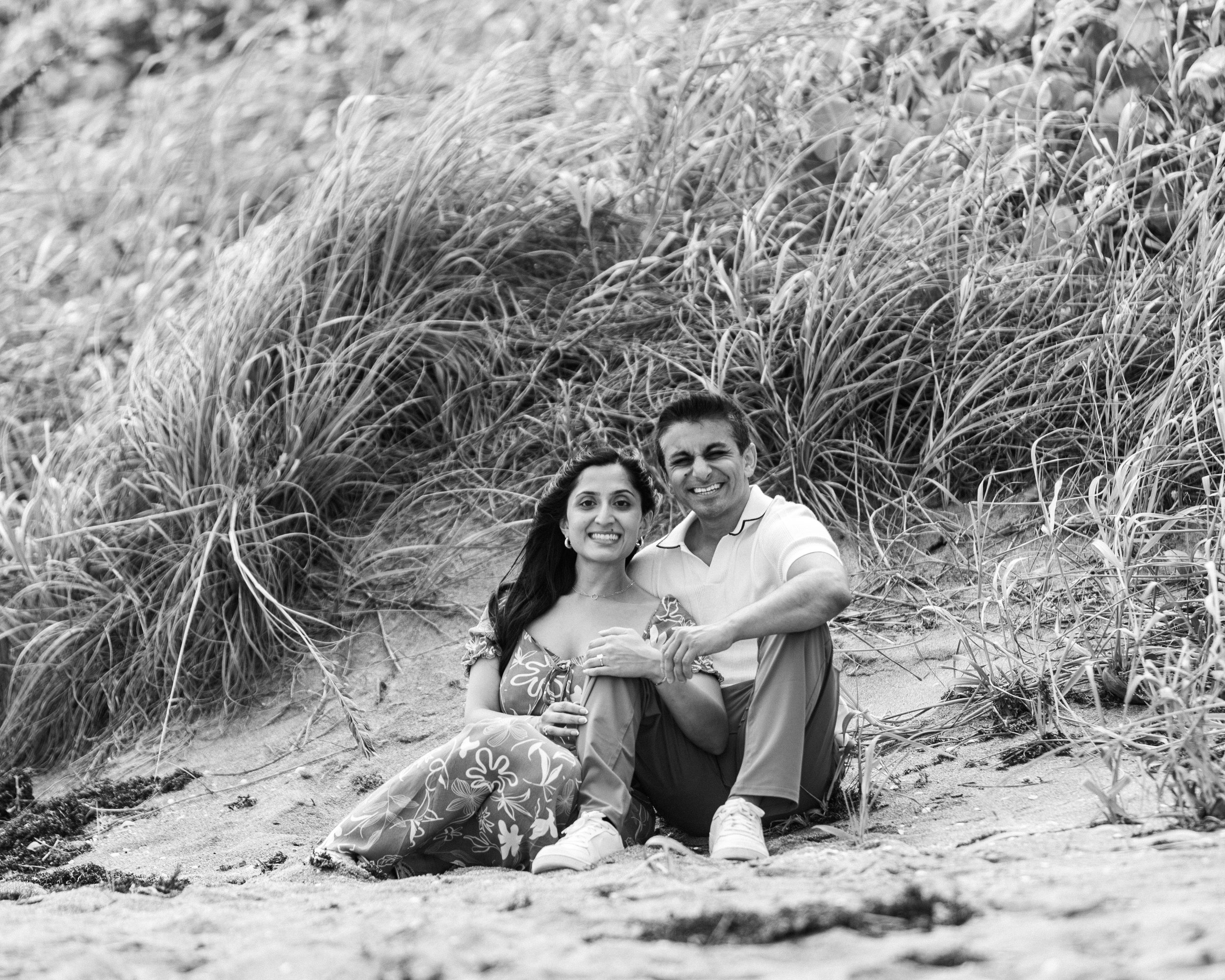 A smiling couple sitting on a sandy beach in front of tall grass and dunes, posing for the photograph.