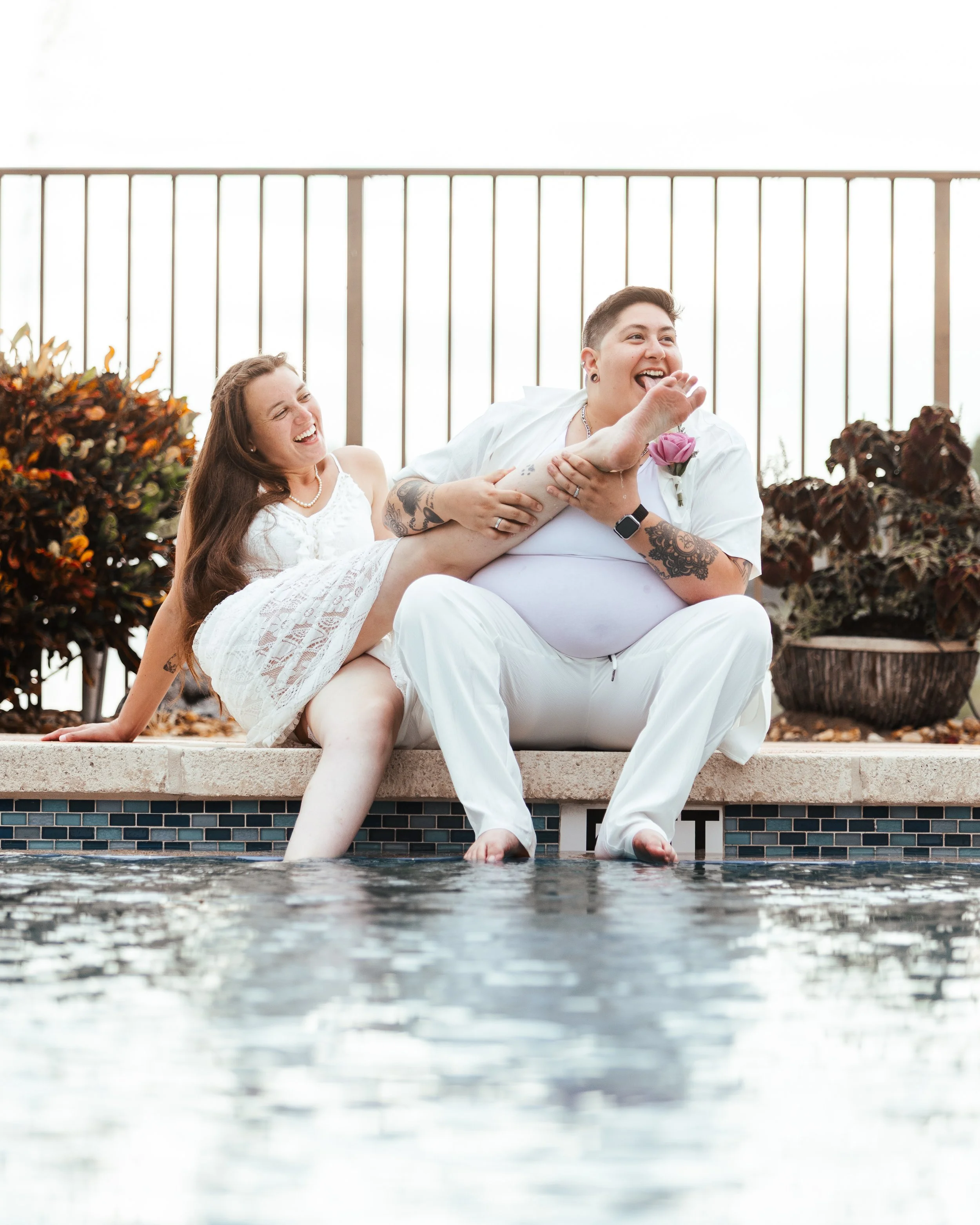 Two people sitting by a pool, one person playfully holding the other's leg, both laughing and enjoying the moment.