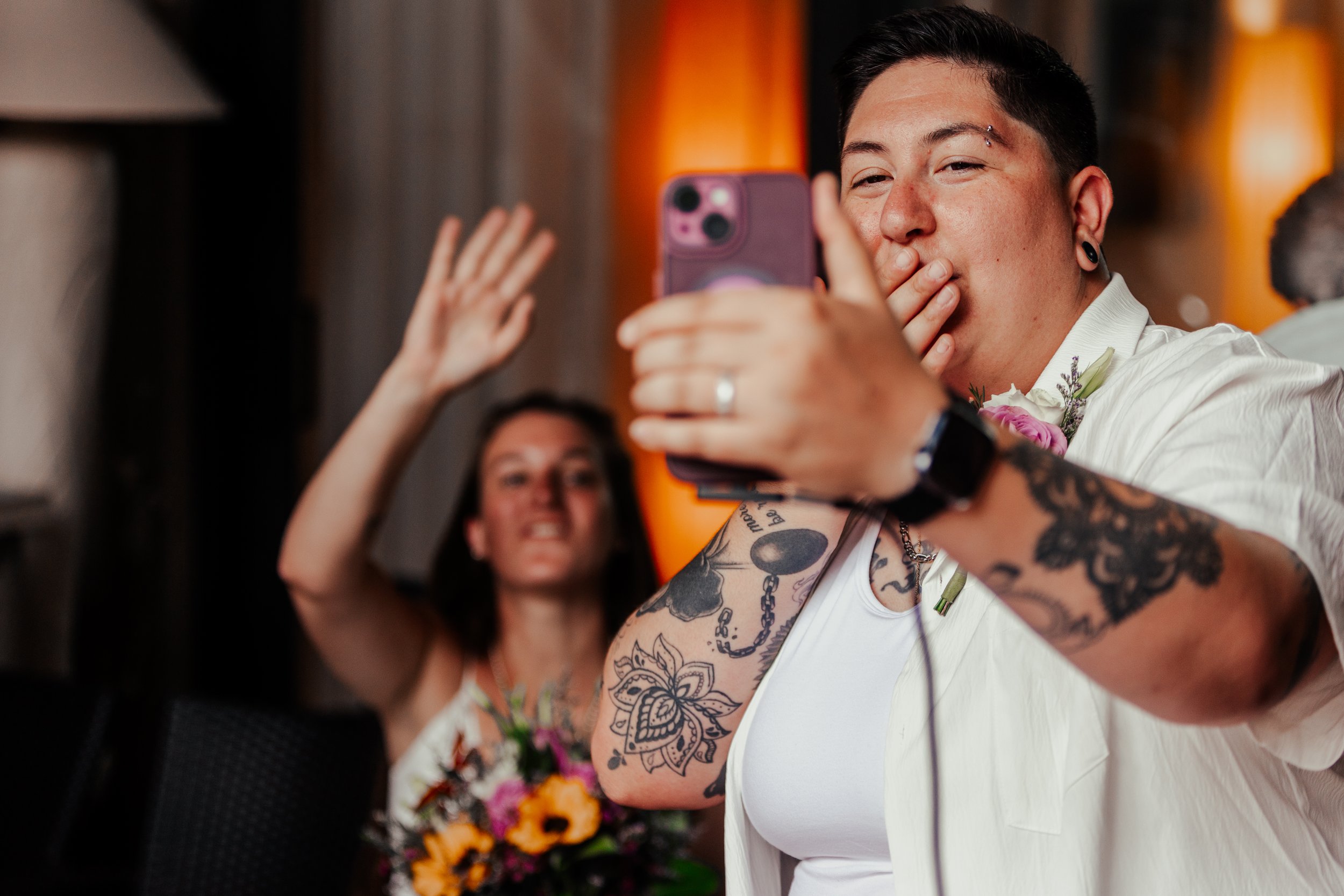 A person with tattoos taking a selfie with another person in the background waving. They are at an indoor event with warm lighting and floral decorations.