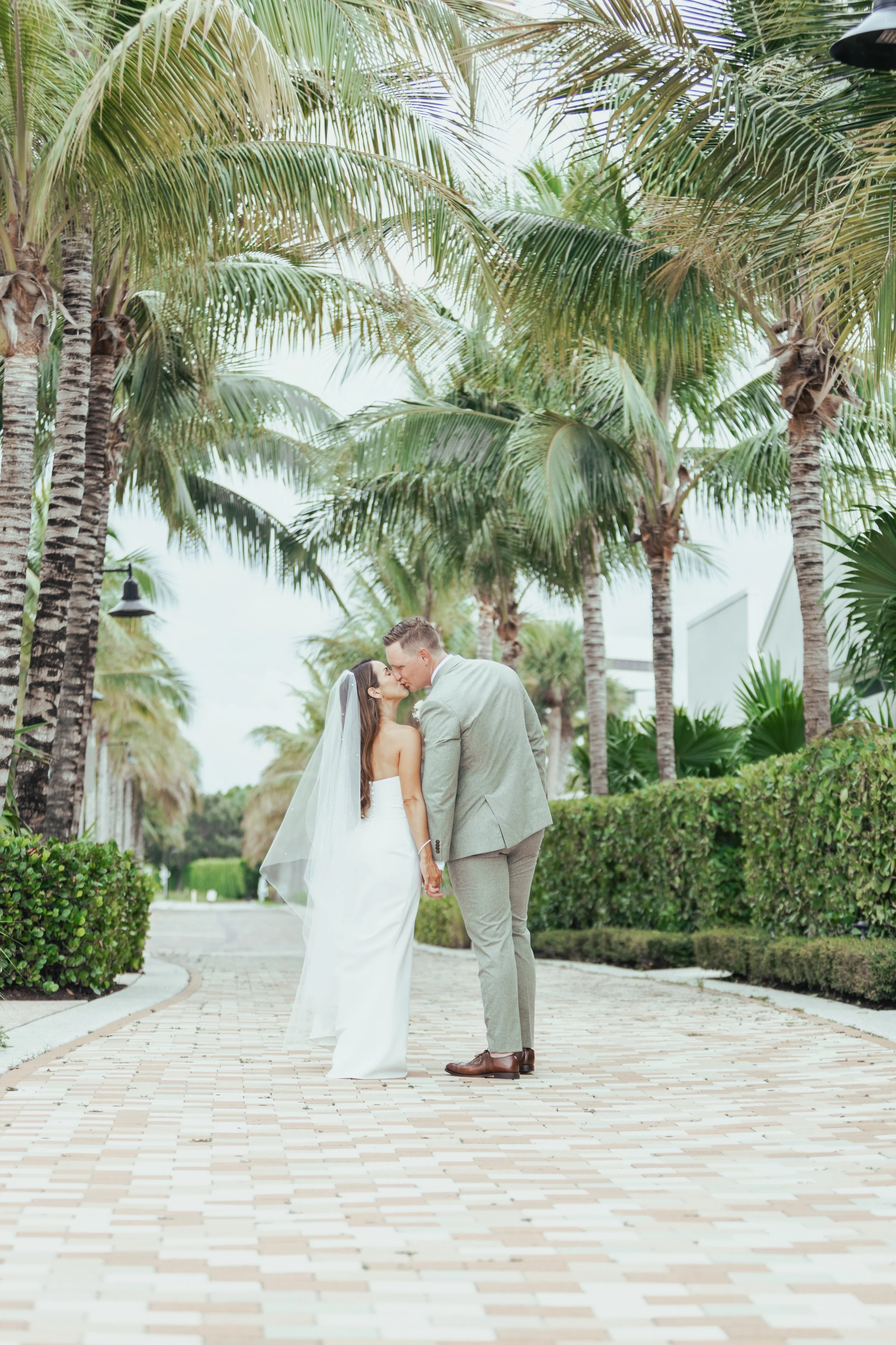 A bride and groom sharing a kiss in a tropical outdoor setting with palm trees and neatly trimmed bushes.