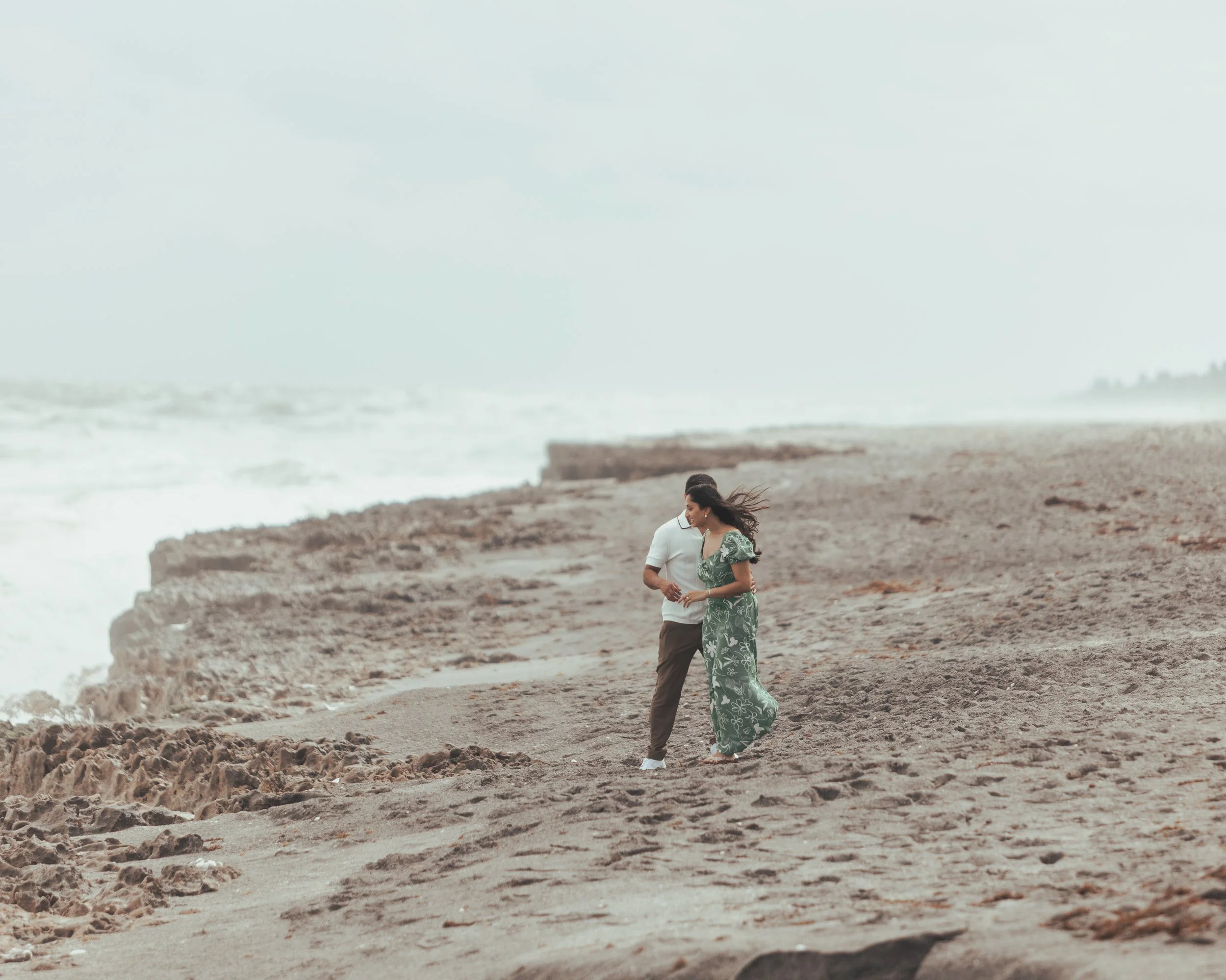 A couple walking on a beach with sandy and rocky terrain, ocean waves in the background, and a cloudy sky.