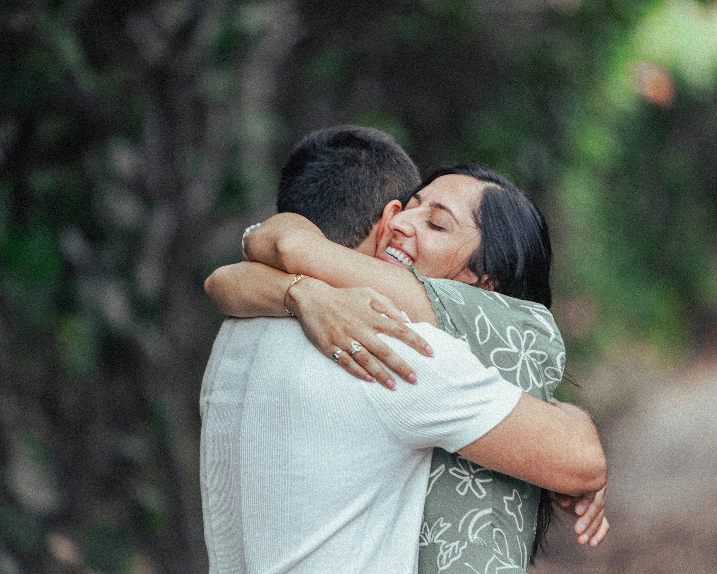 A woman and a man are embracing outdoors, smiling, with the woman’s eyes closed and arms around the man's neck.