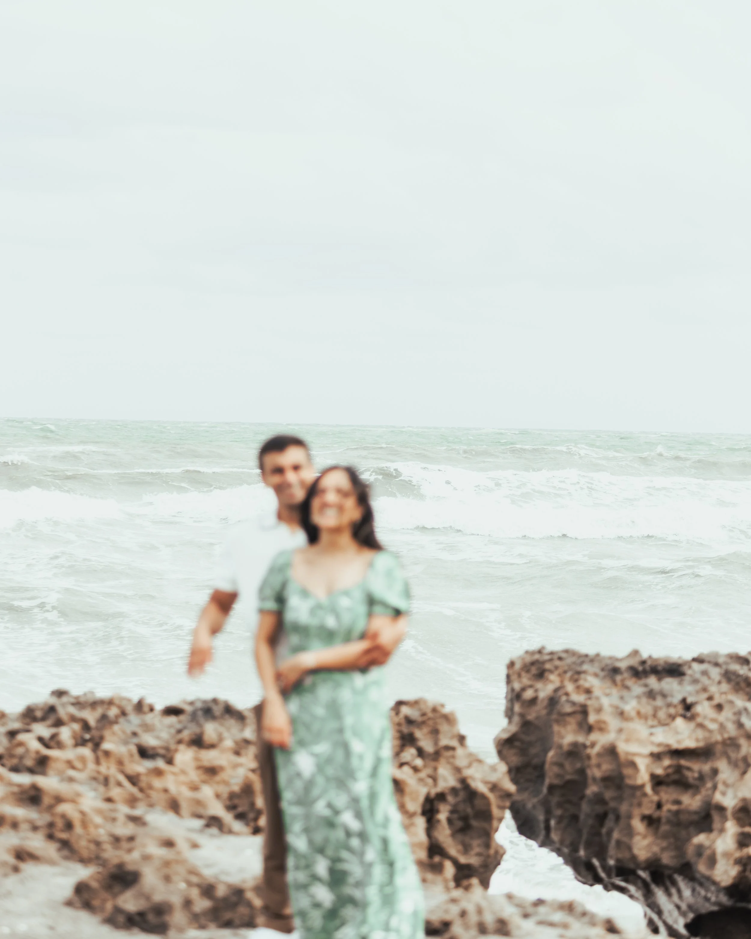A blurred photo of a smiling couple standing on rocks at the beach, with the ocean and cloudy sky in the background.