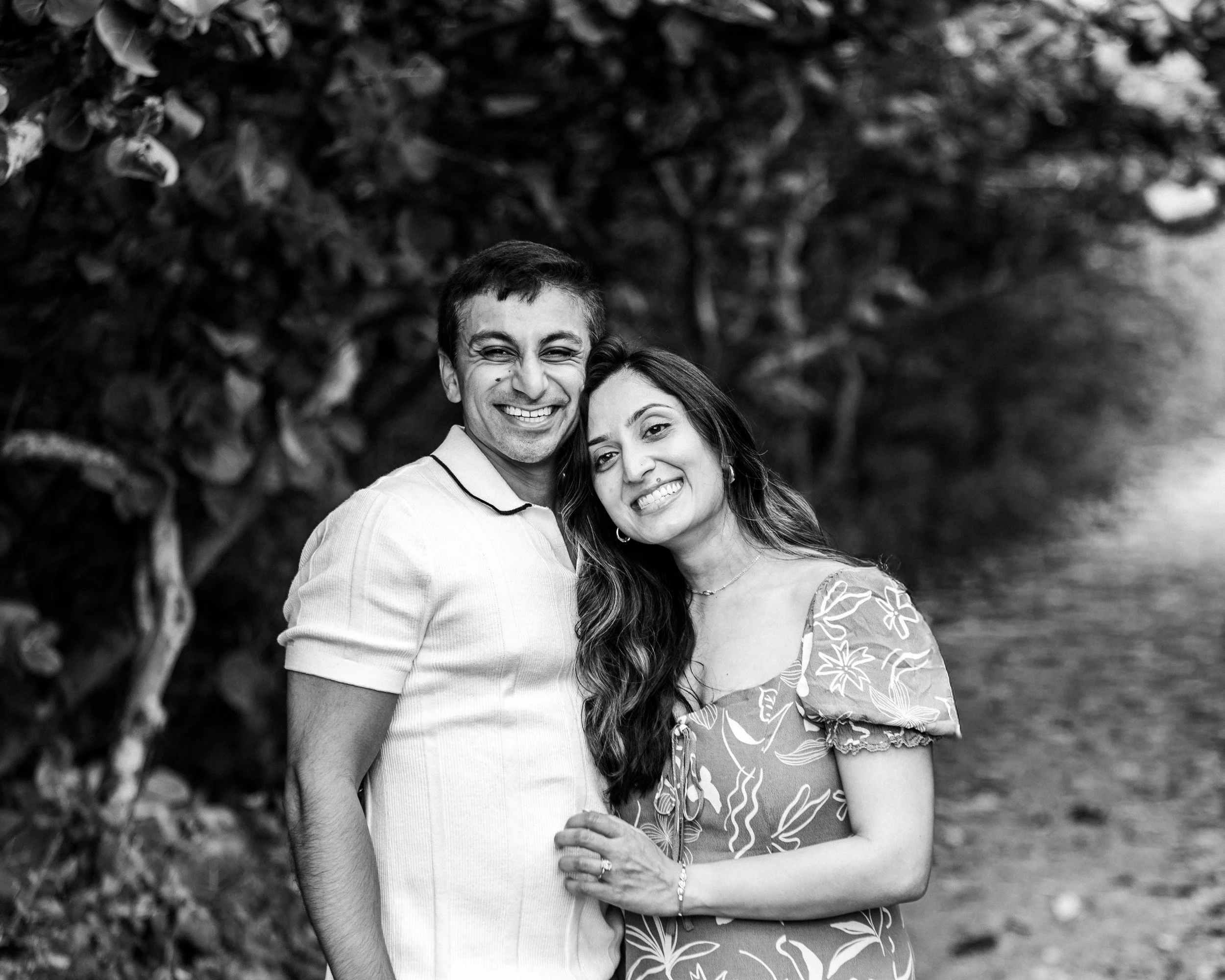 A black and white photo of a smiling couple standing outdoors in front of a tree. The man is wearing a short-sleeved polo shirt, and the woman is wearing a floral dress with puffed sleeves. They are close together, with the woman's hand resting on th