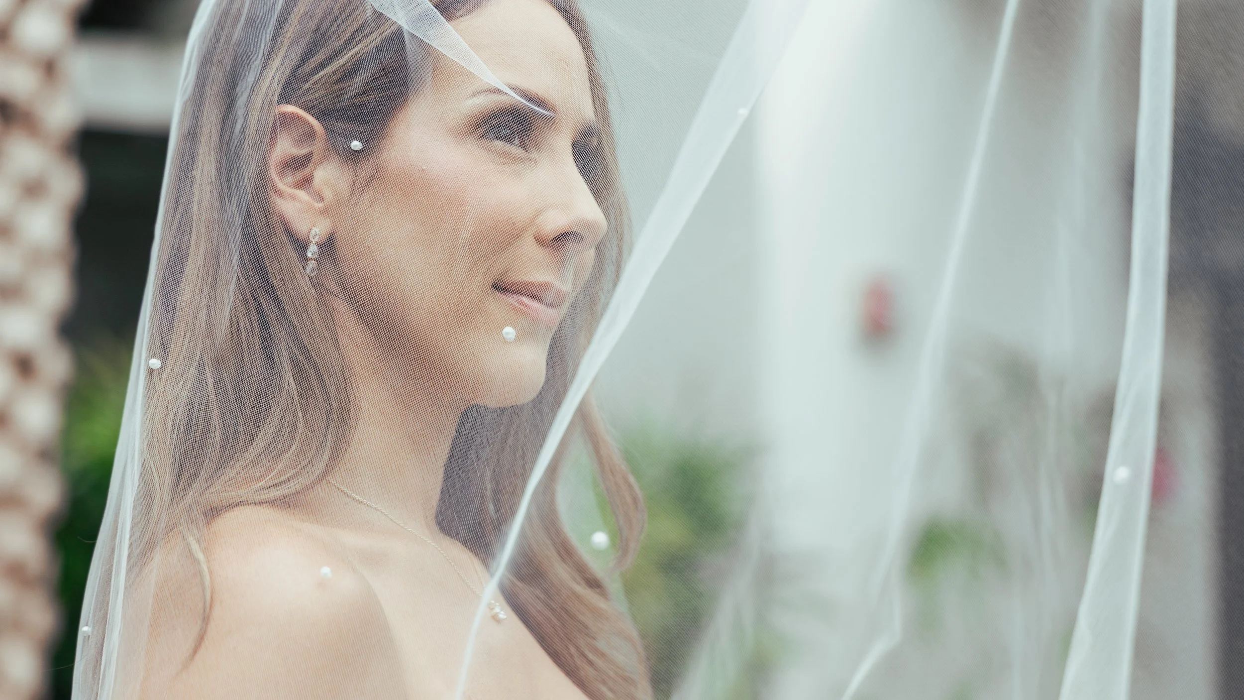 A bride with earrings and a necklace, smiling softly, is partially covered by a sheer veil. She is outdoors with a blurred brick wall and greenery in the background.