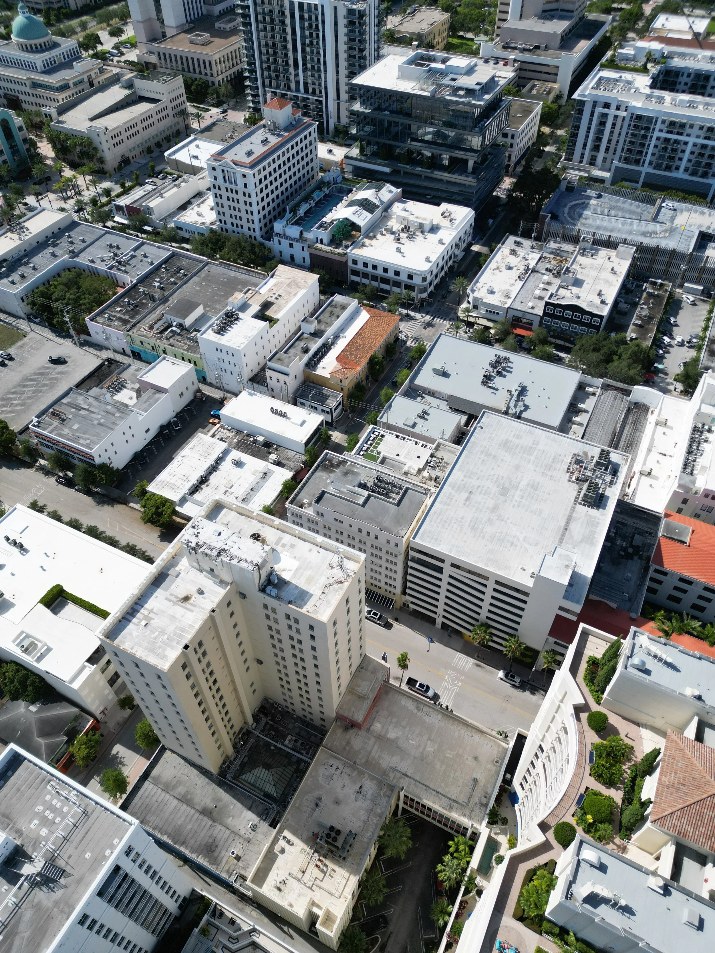 Aerial view of a city with high-rise buildings, office buildings, commercial structures, and a grid of streets with cars, surrounded by trees and green spaces.