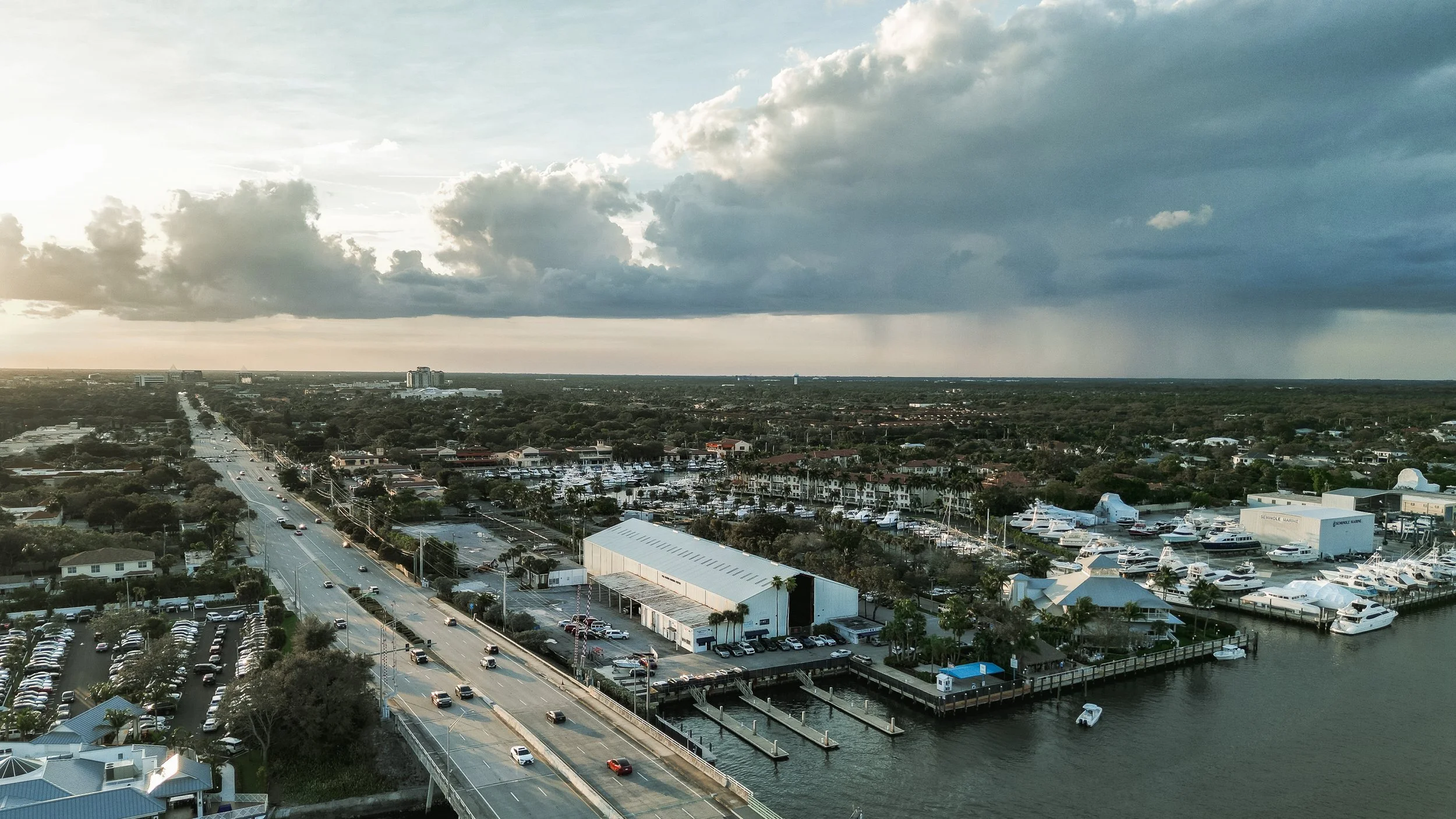 Aerial view of a coastal city with a marina filled with yachts, a bridge crossing a waterway, a main road with cars, residential and commercial buildings, and a cloudy sky overhead.