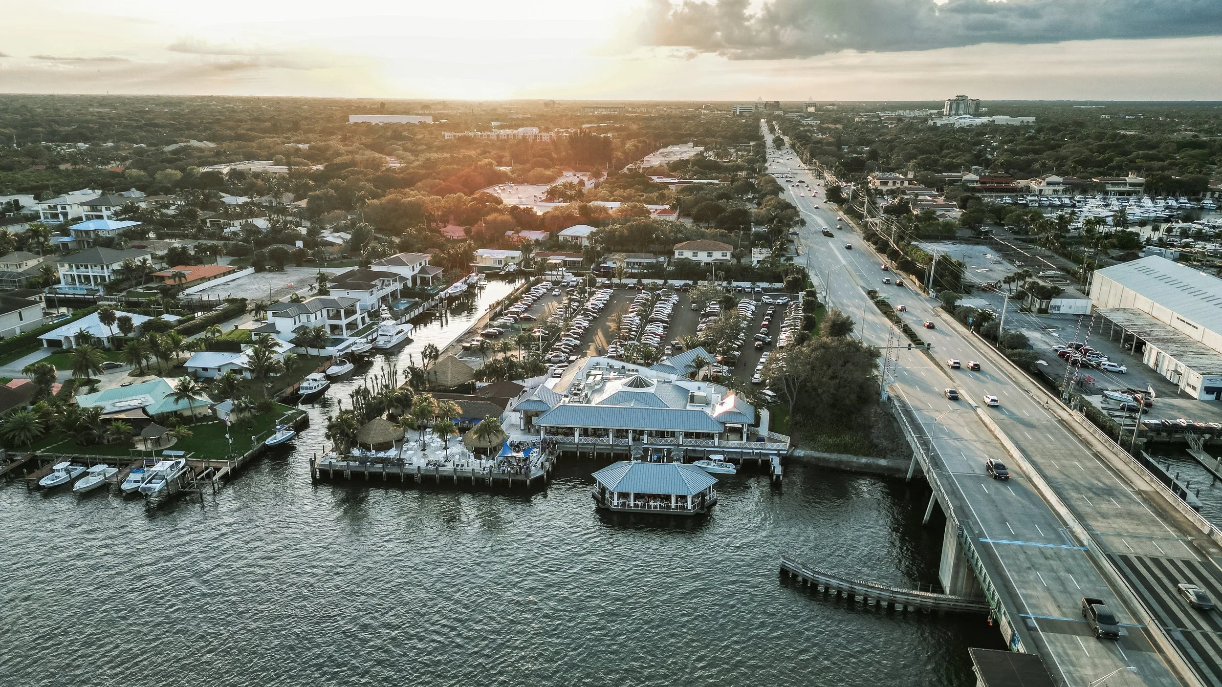 Aerial view of a coastal city area showing houses, a marina with boats, parking lots, a river, a bridge, and a road with moving cars under a cloudy sky.