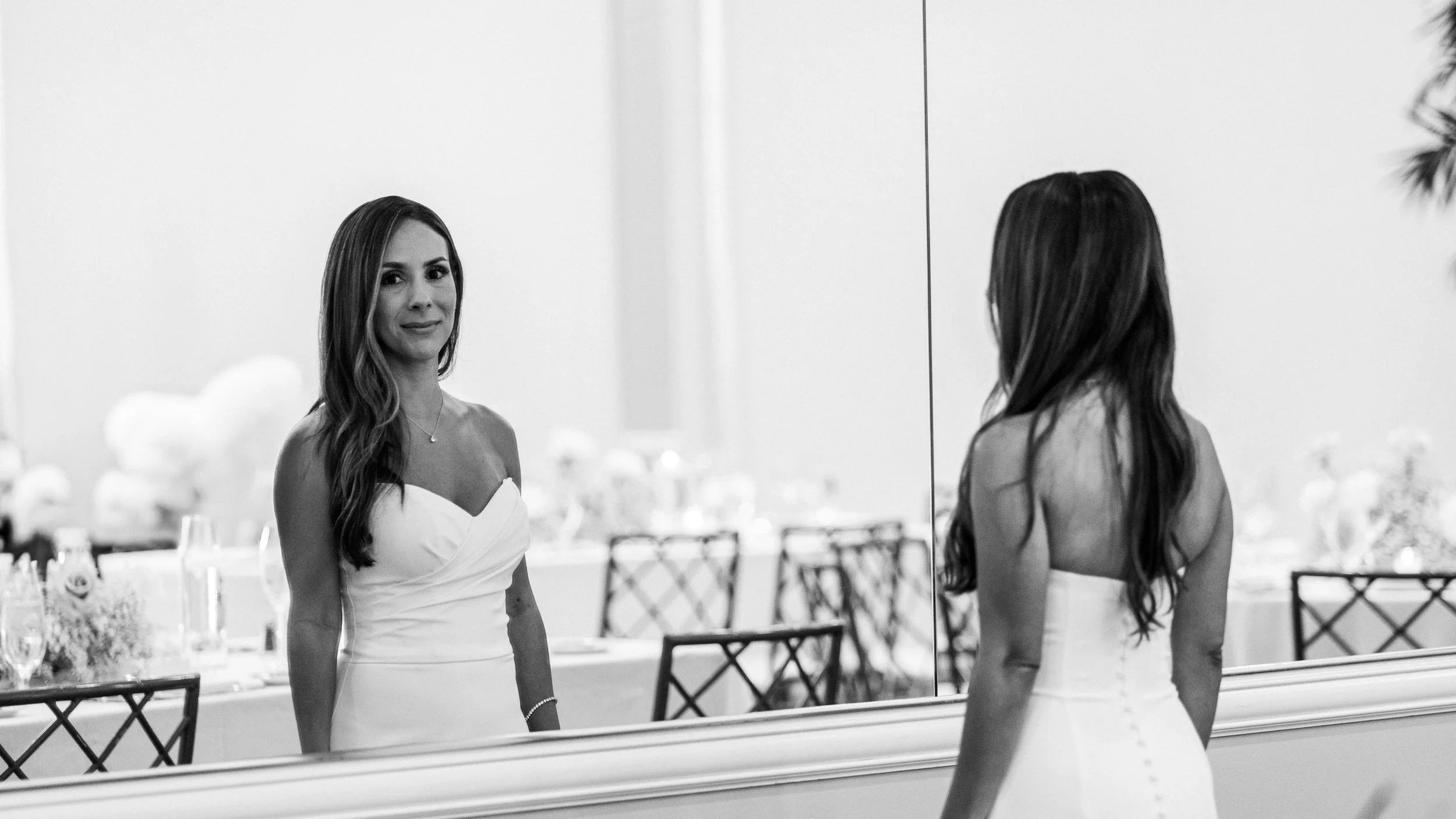 A woman in a white strapless dress looking at her reflection in a mirror, with a decorated table and chairs in the background.