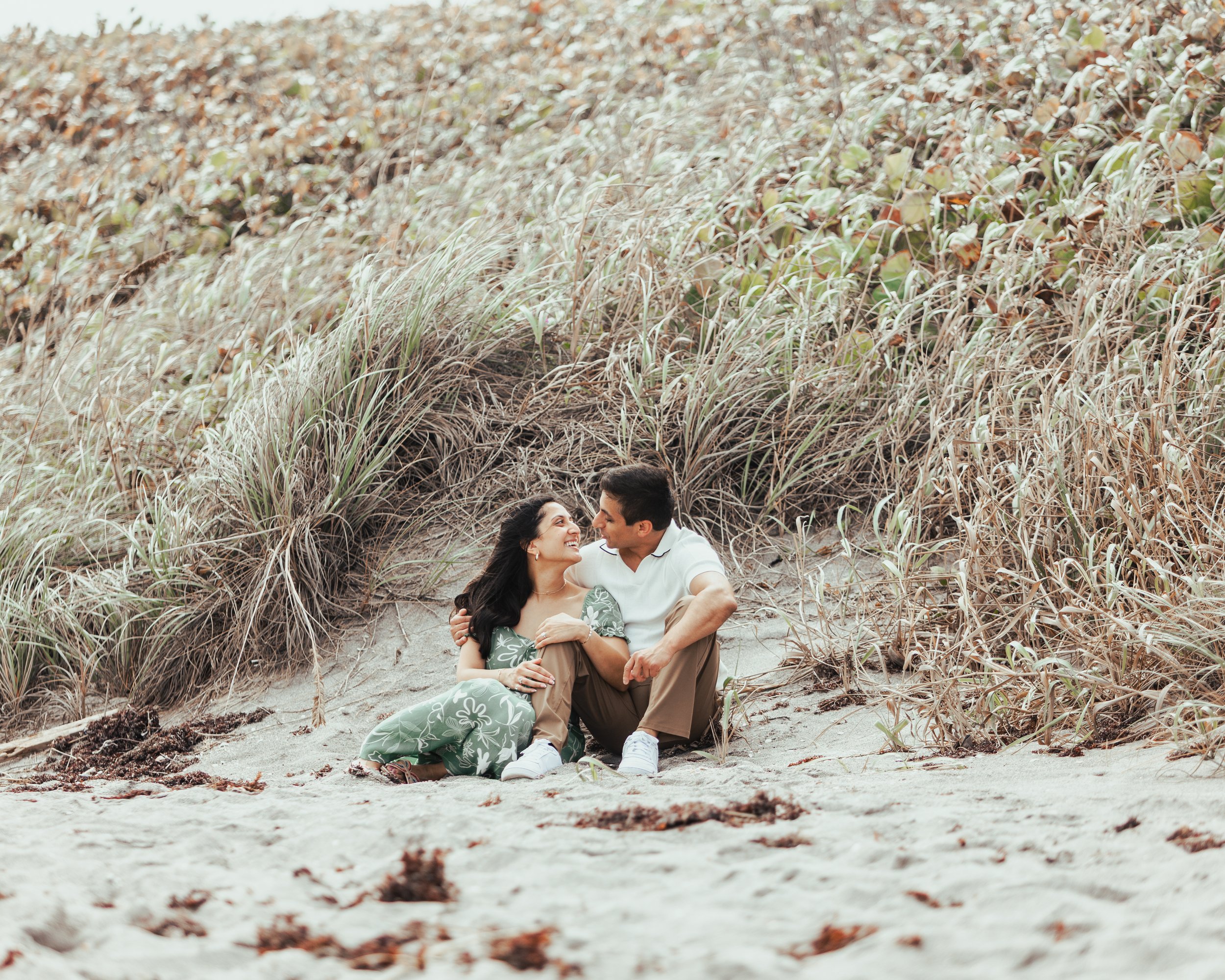 A couple sitting on the sand in front of tall grasses, smiling and looking into each other's eyes at a beach.