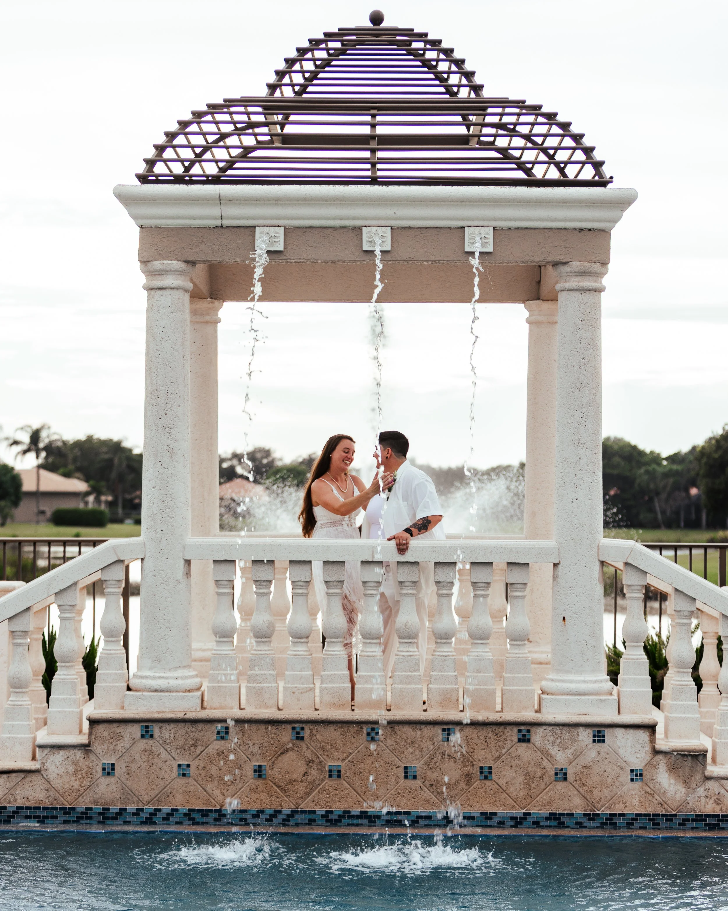 A couple standing under a fountain gazebo in a pool, smiling and touching for a romantic moment.