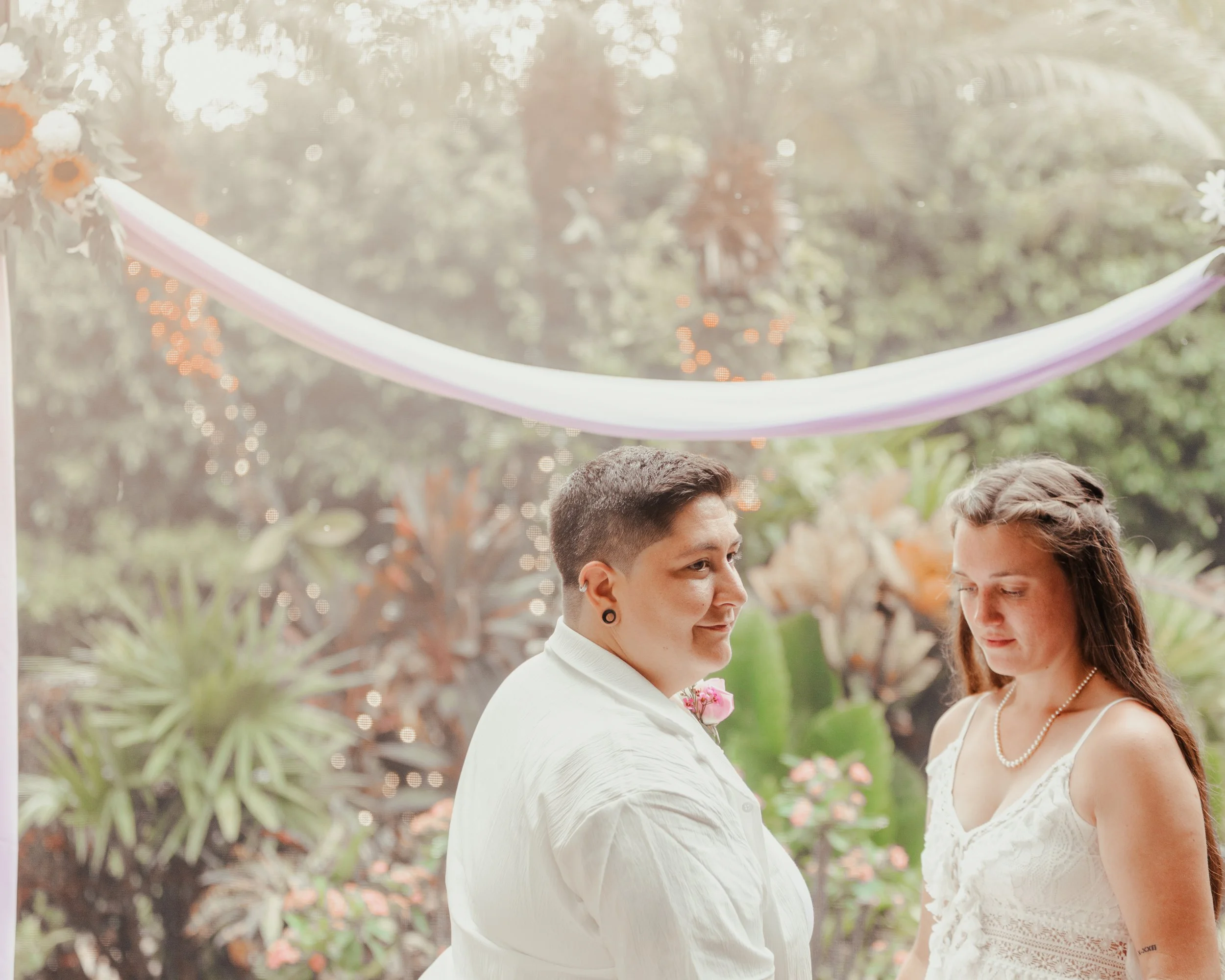 A same-sex couple standing together outdoors during a wedding ceremony, with decorative string lights and greenery in the background.