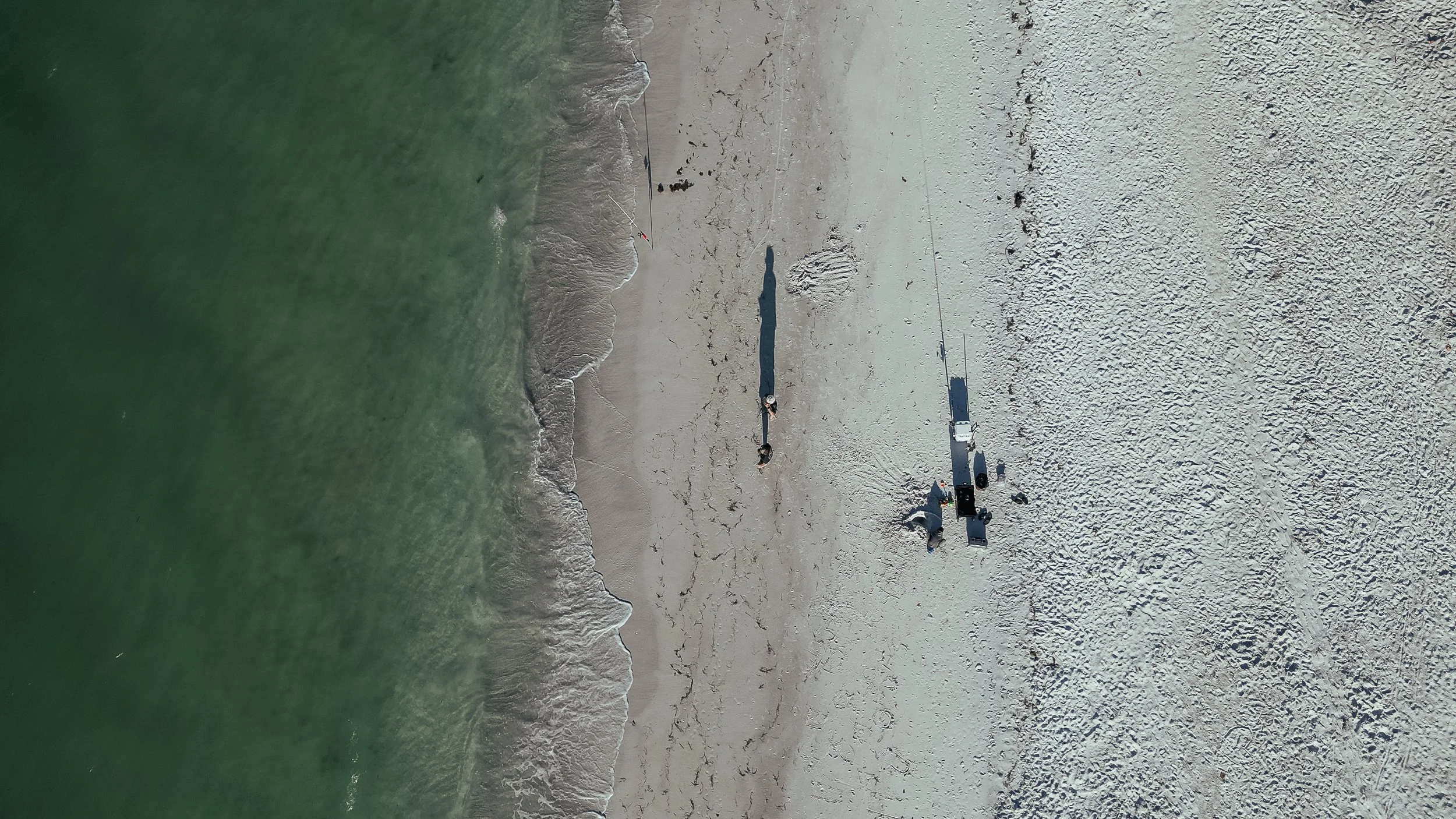 Overhead view of a beach with waves on the left and a sandy shore on the right, with people and equipment set up on the sand.