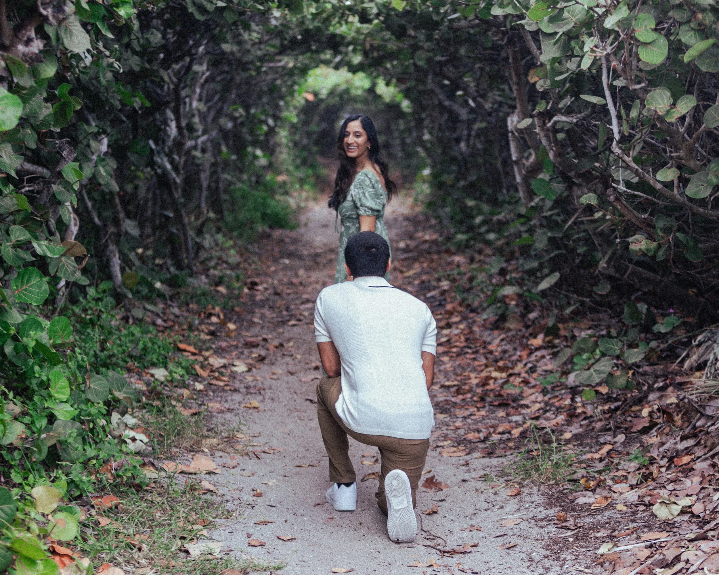 A man is kneeling on one knee in front of a smiling woman on a nature trail surrounded by dense greenery.