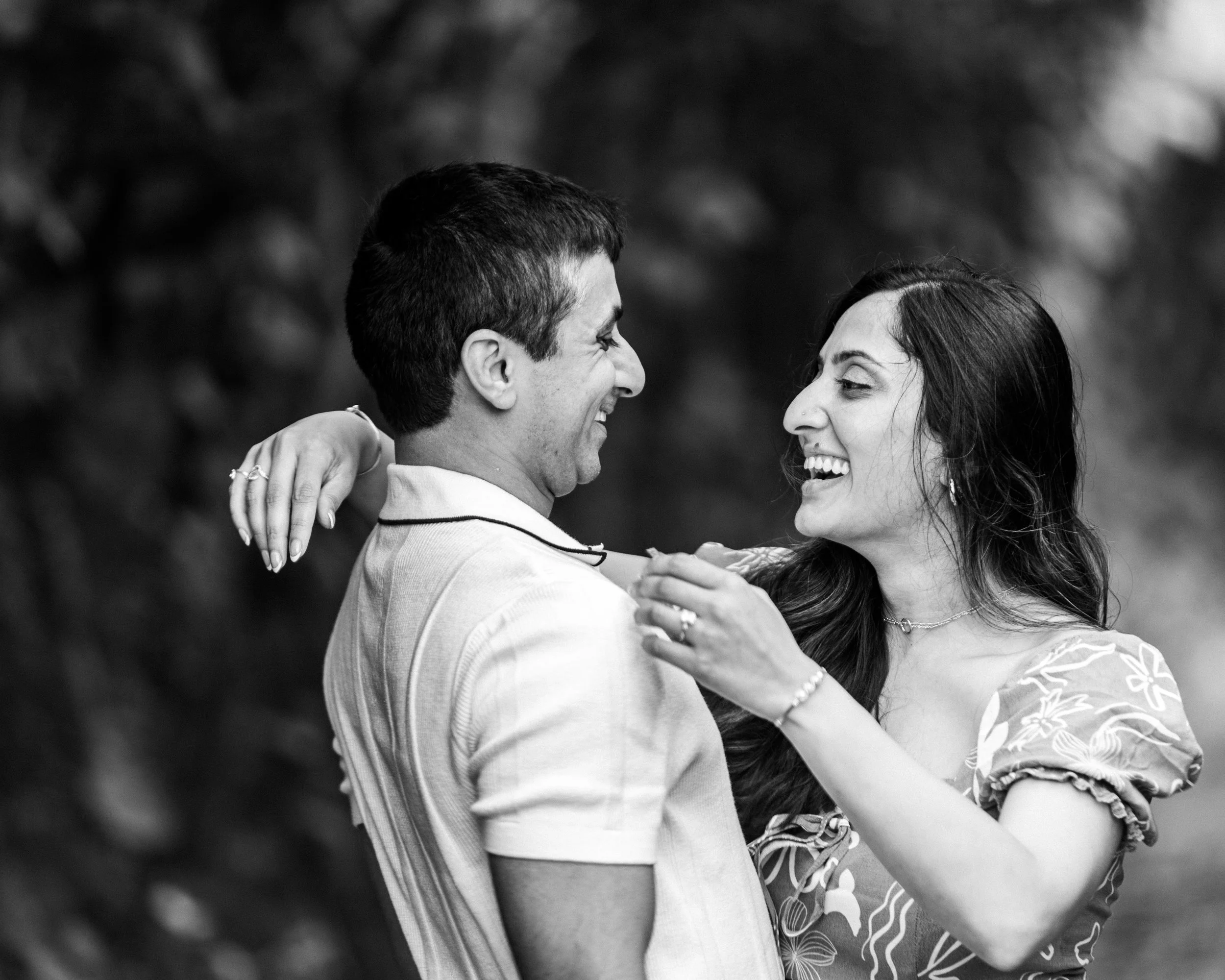 A couple smiling and gazing at each other outdoors, with the woman holding the man's collar, in a candid black and white photo.
