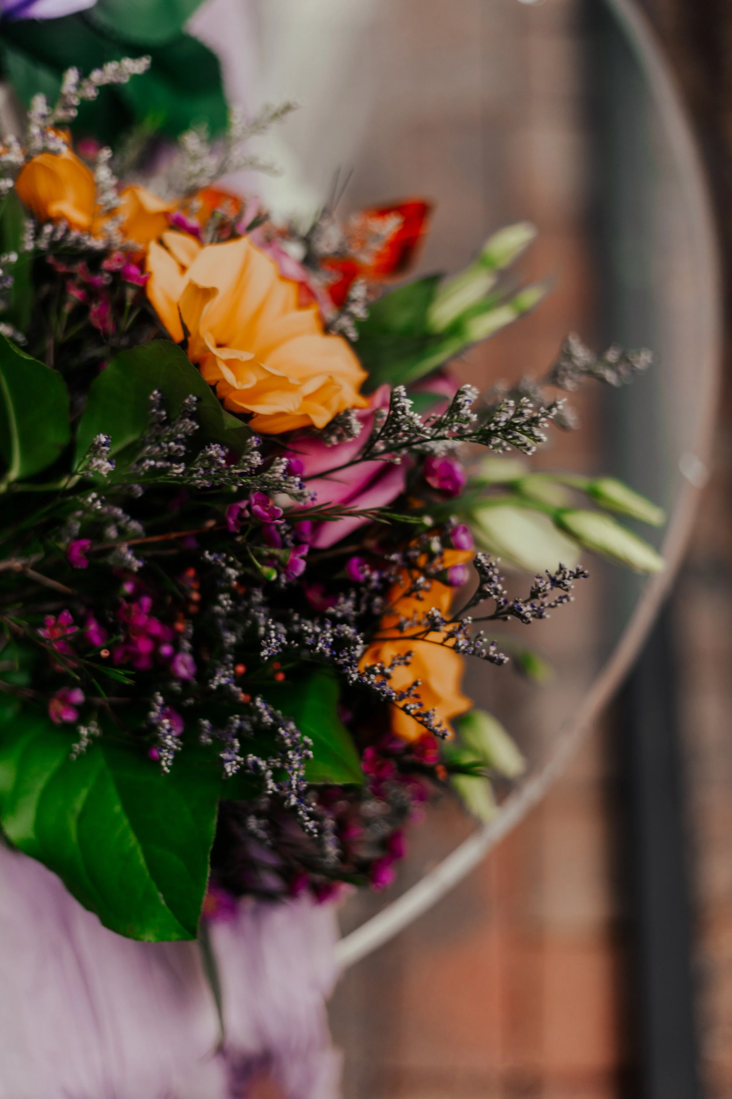 Close-up of a colorful flower arrangement with yellow, pink, and purple flowers and green leaves, with a brick wall background.