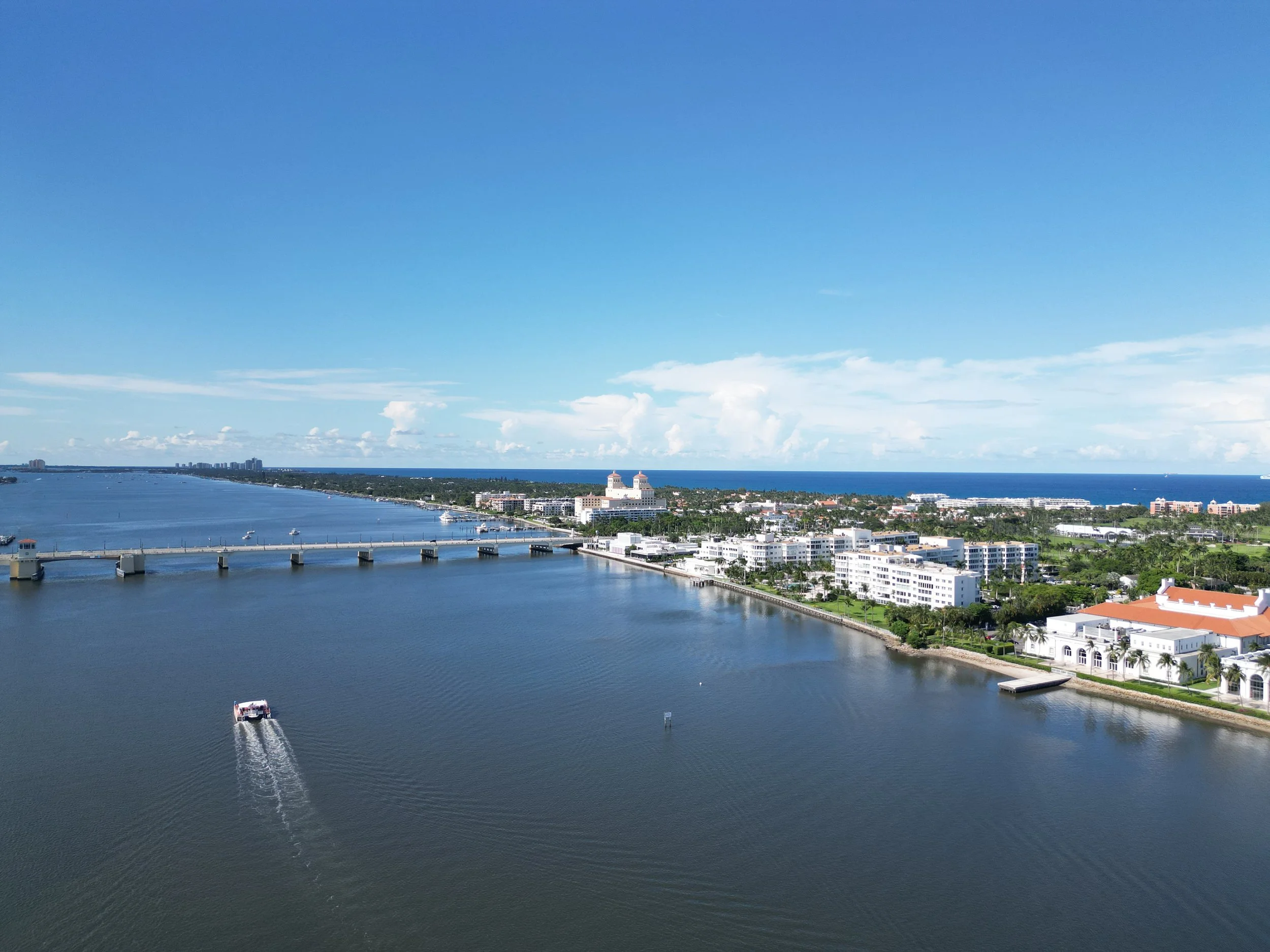 Aerial view of a coastal city with a river or canal, a bridge, white buildings, and the ocean in the background under a blue sky with clouds.
