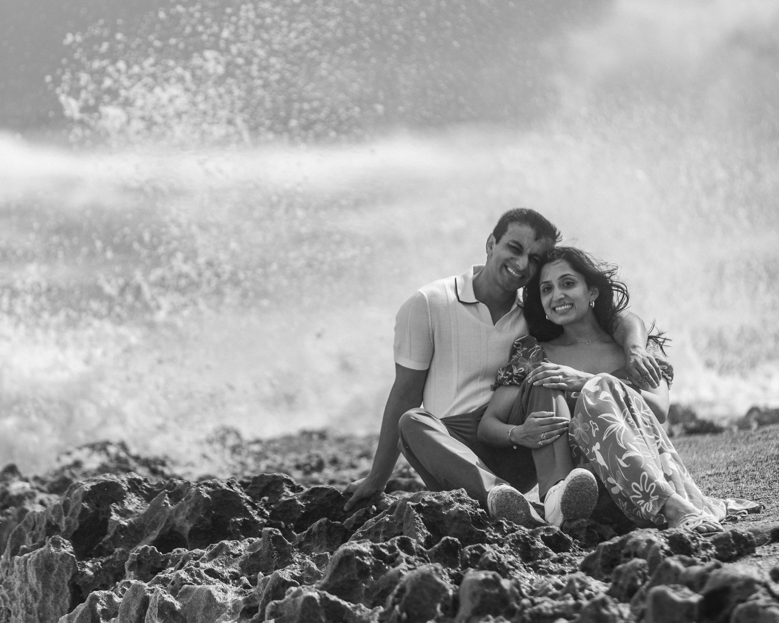 A black and white photo of a smiling man and woman sitting on rocks by the ocean, kissed by splashing waves, embracing and looking at the camera.