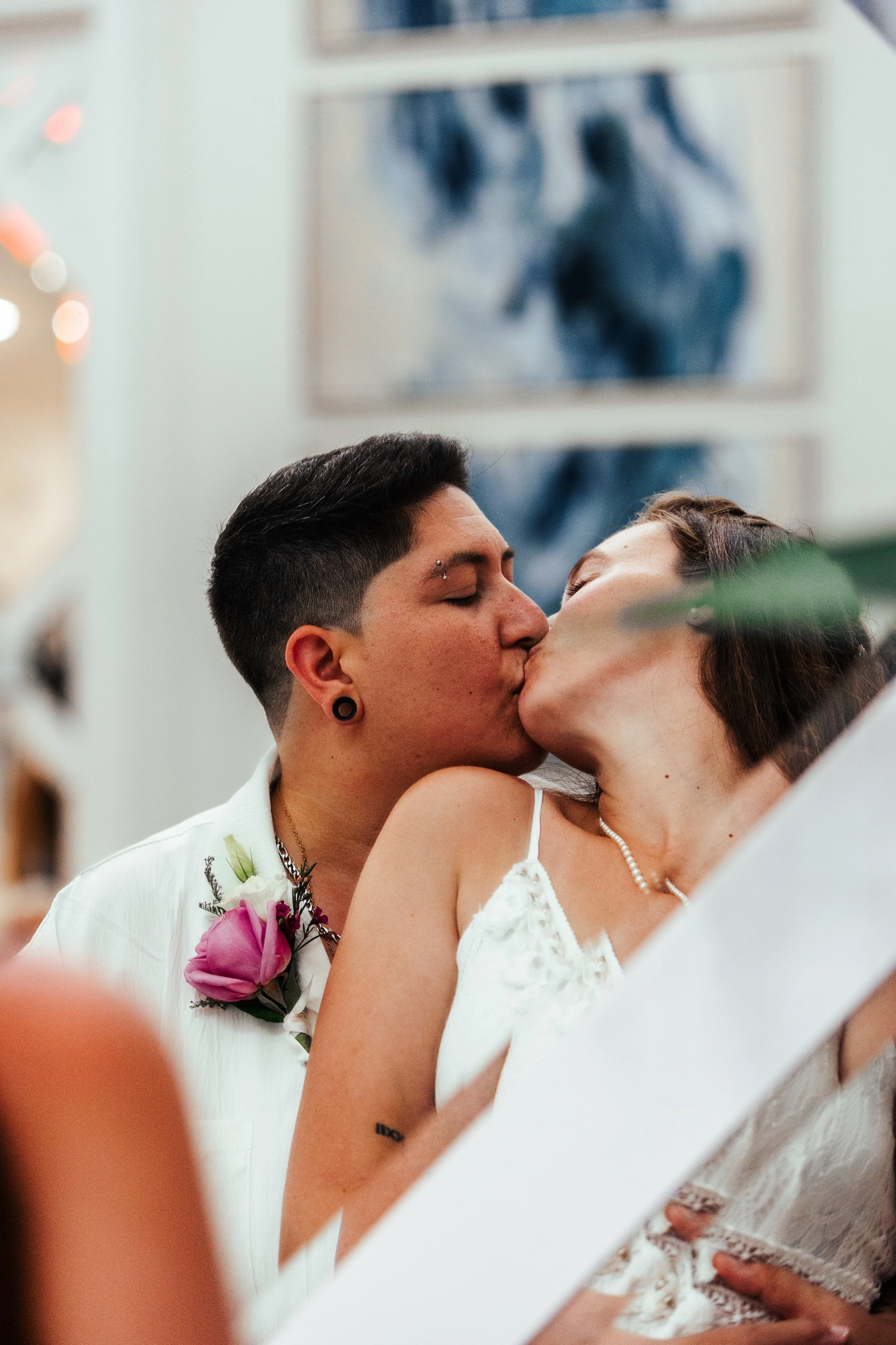 Two women kissing at a celebration, one with short black hair and piercings, the other with short brown hair, wearing a white dress and pearl necklace.