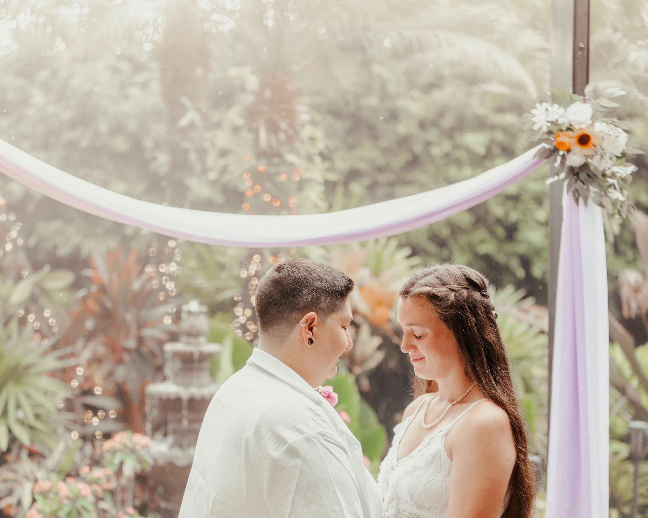 A couple getting married outdoors under a decorated arch with purple fabric and flowers, with trees and string lights in the background.