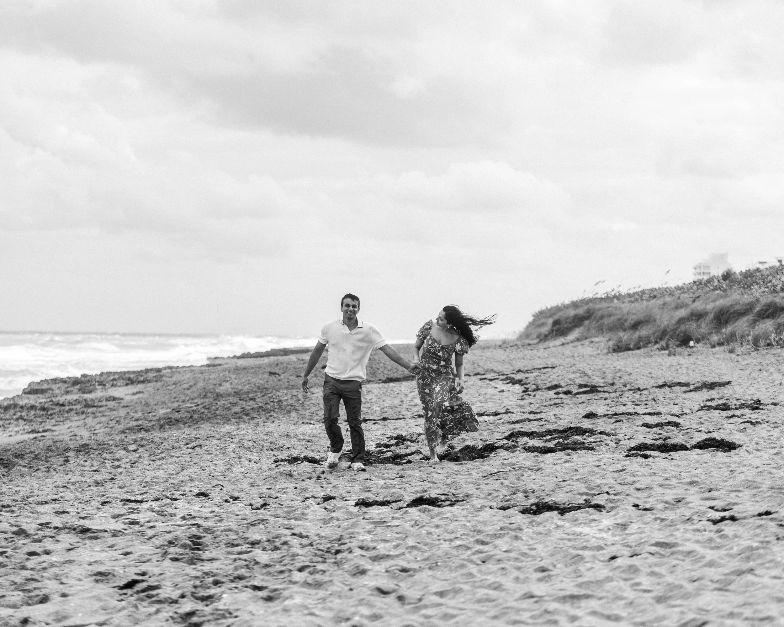 A couple walking hand in hand on a beach, with the woman looking at the man and the man smiling, in a black and white photograph.