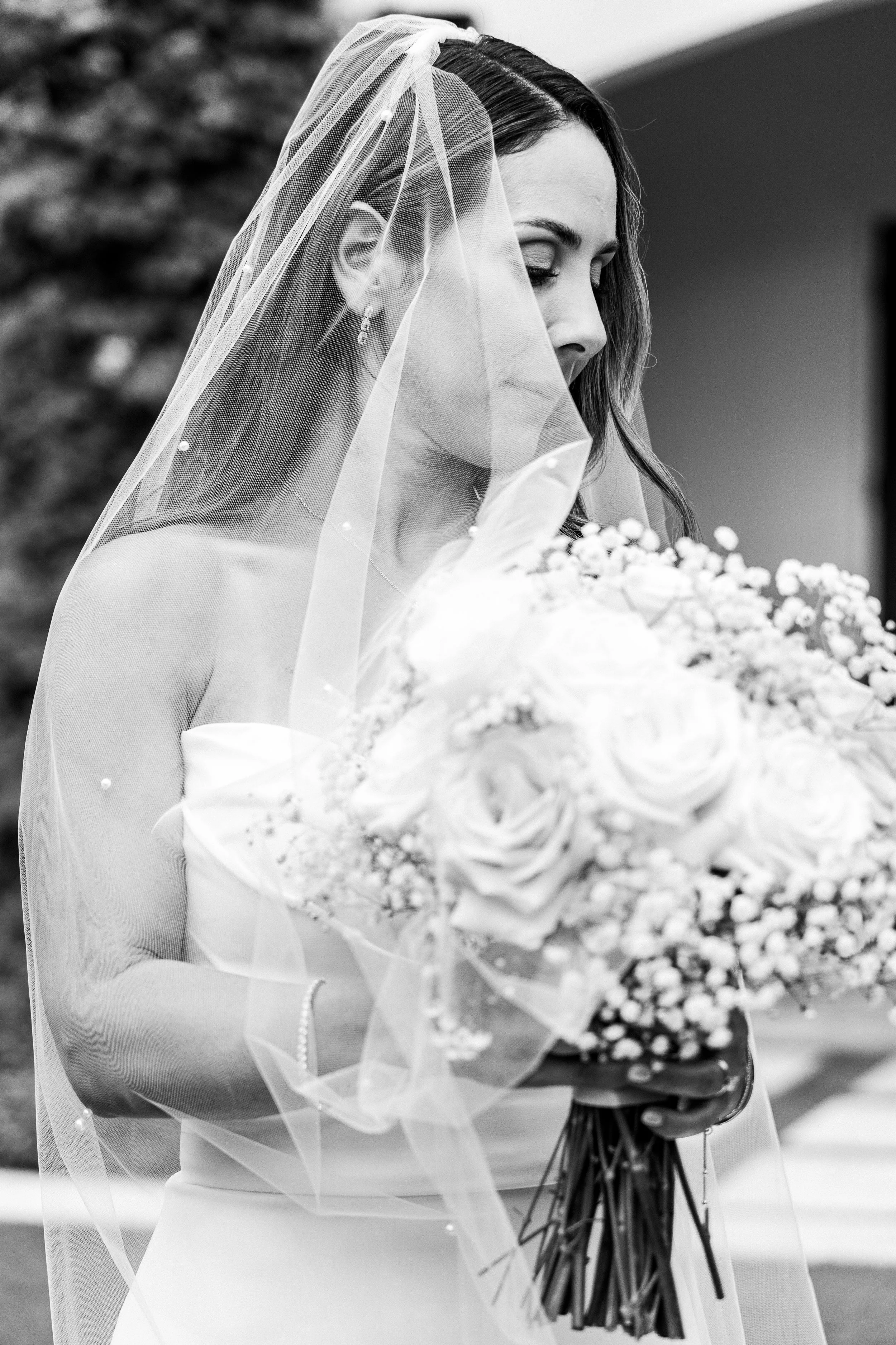 A bride with her eyes closed, wearing a strapless wedding gown and veil, holding a bouquet of roses and baby's breath.