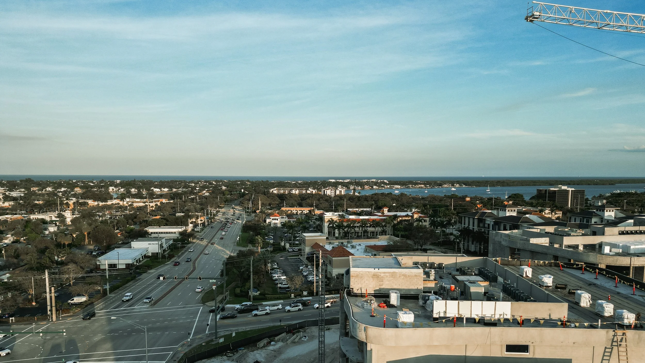 Cityscape view from a high vantage point showing streets with cars, a mix of commercial and residential buildings, and a body of water in the distance under a blue sky with some clouds.
