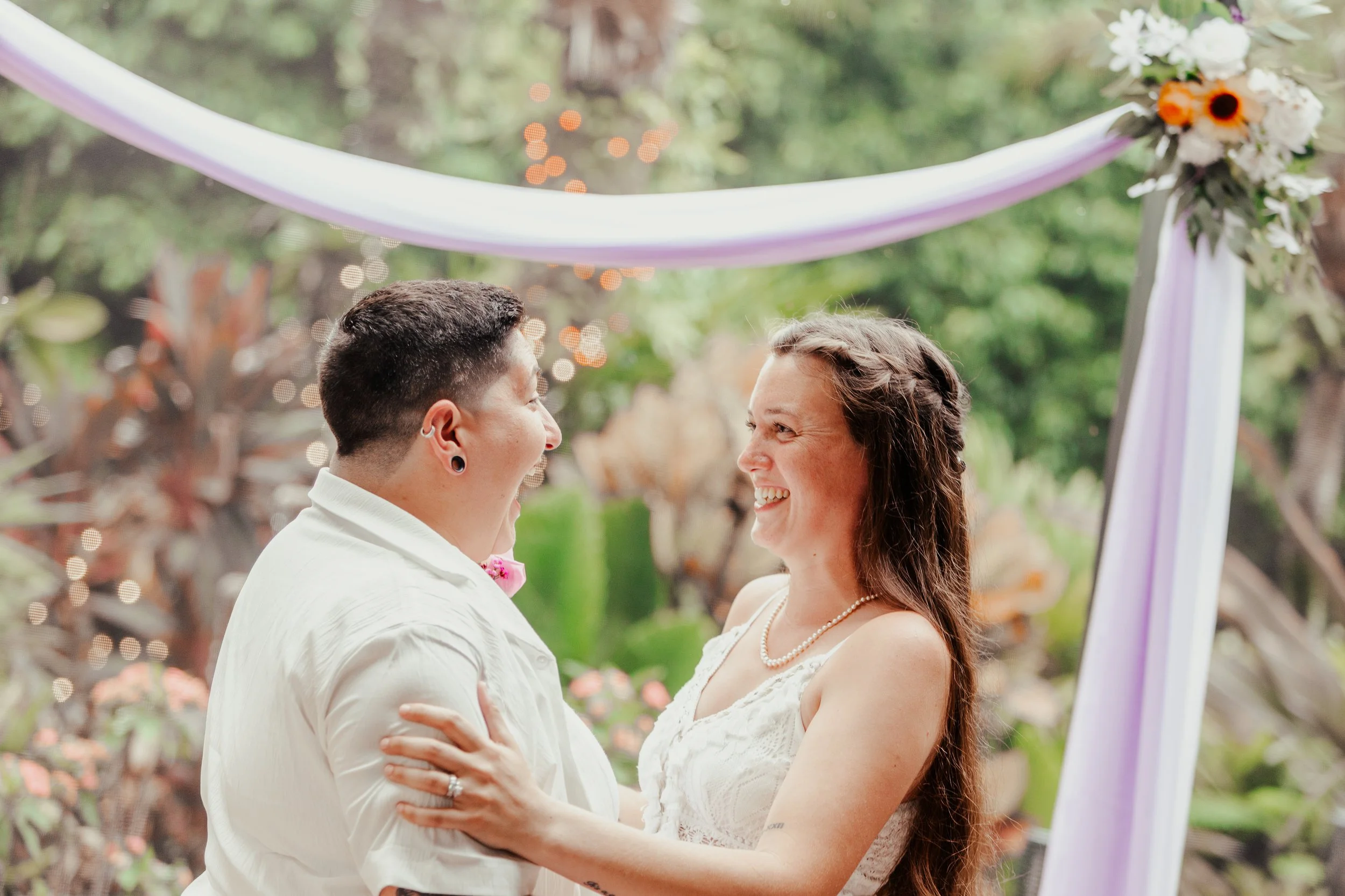 A couple happily getting married outdoors under a decorated archway with purple ribbons and flowers, surrounded by lush greenery.