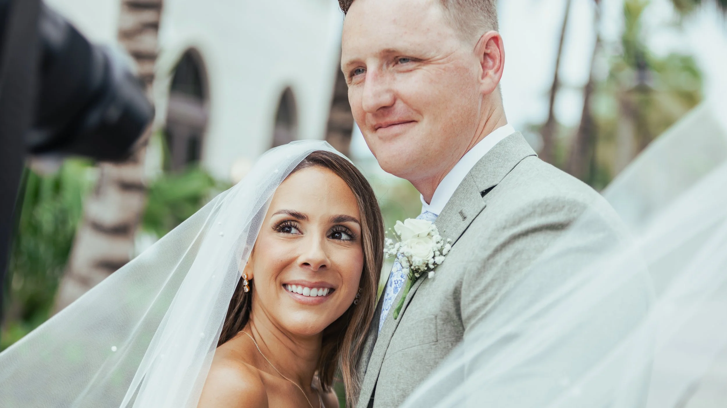Happy bride and groom at their wedding ceremony outdoors, smiling and looking at the officiant, with a wedding veil and boutonniere visible.
