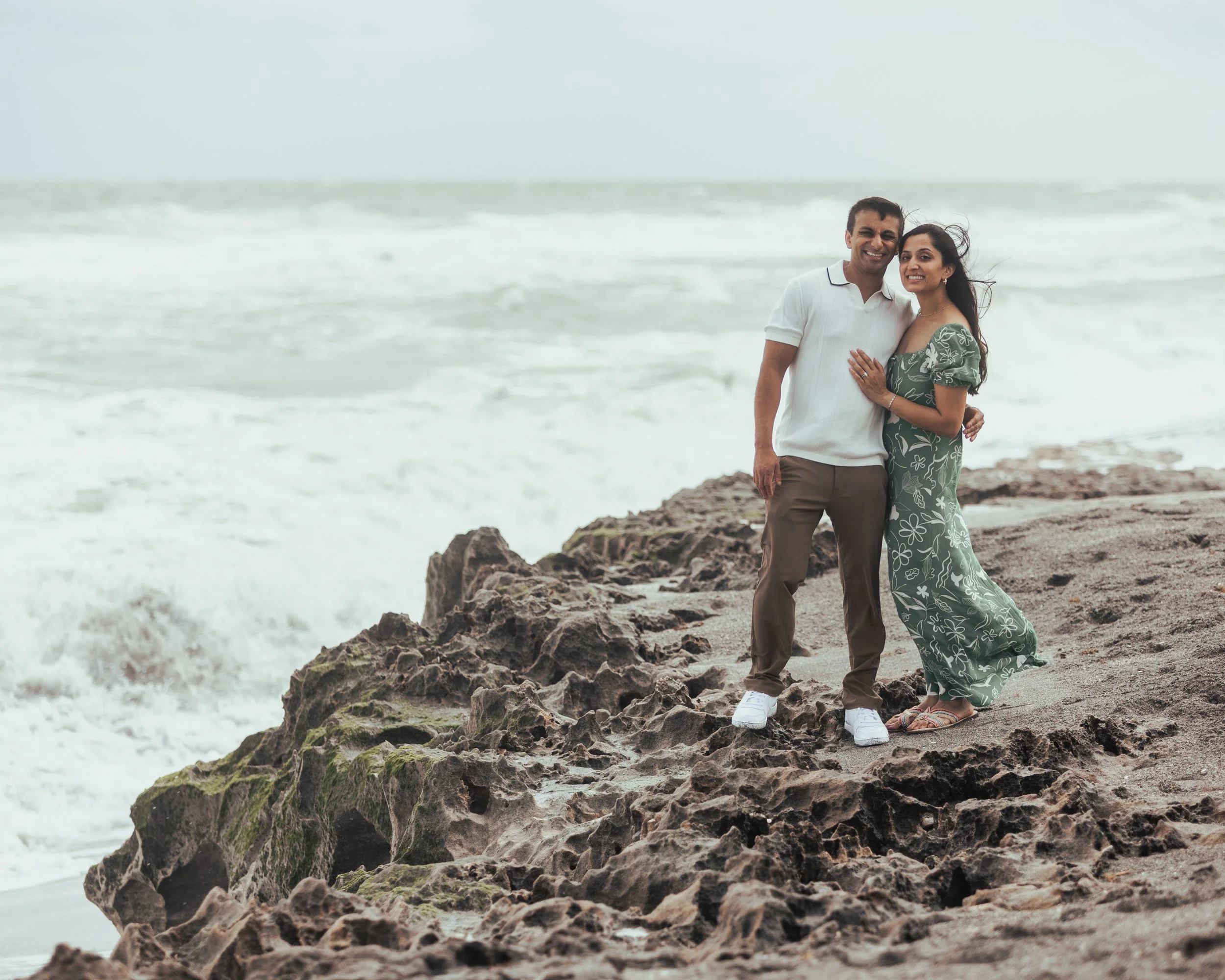 A couple standing on a rocky beach near the ocean, smiling and embracing each other.