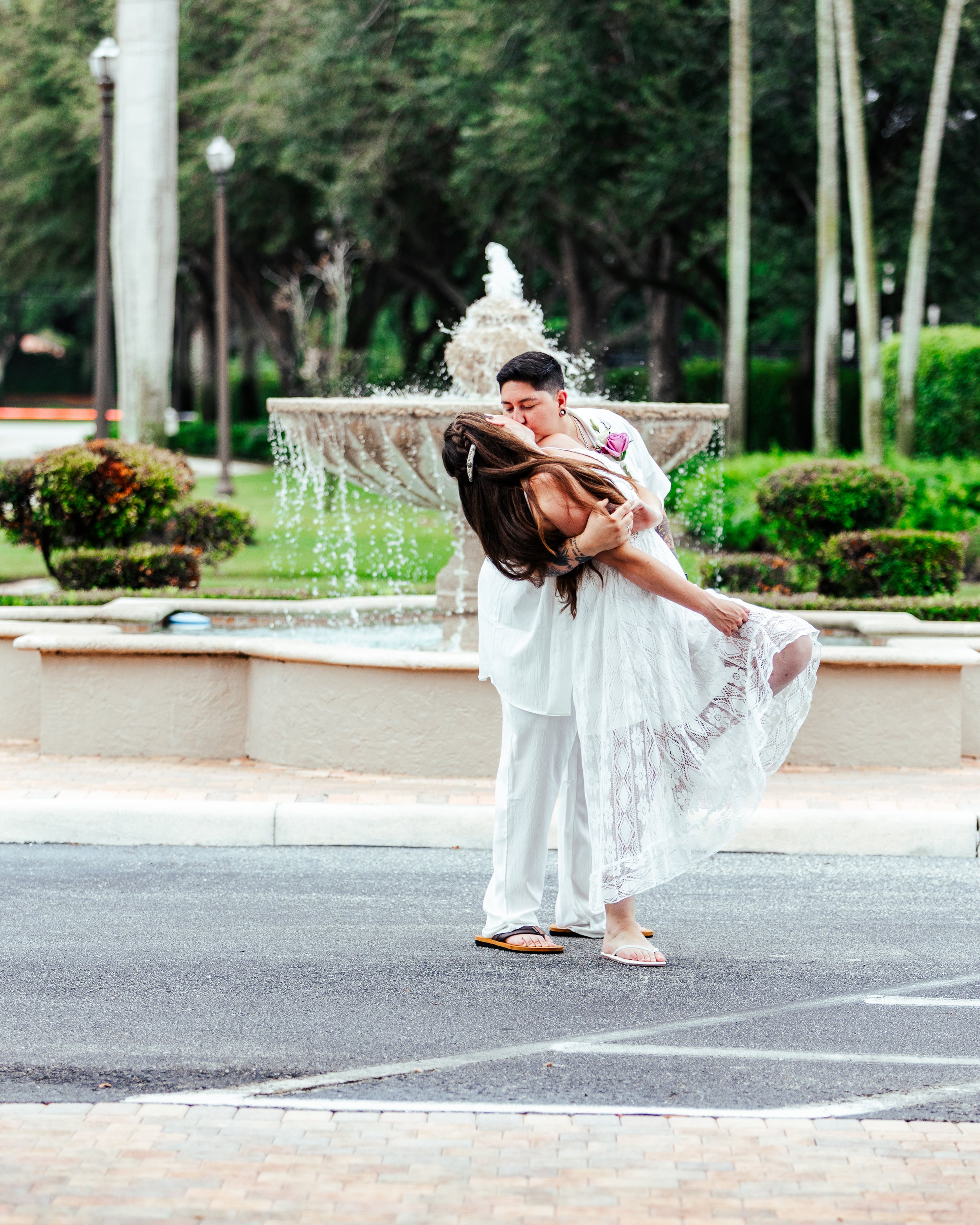 A couple dressed in white, with the man in slider sandals, kiss in front of a fountain in a park.