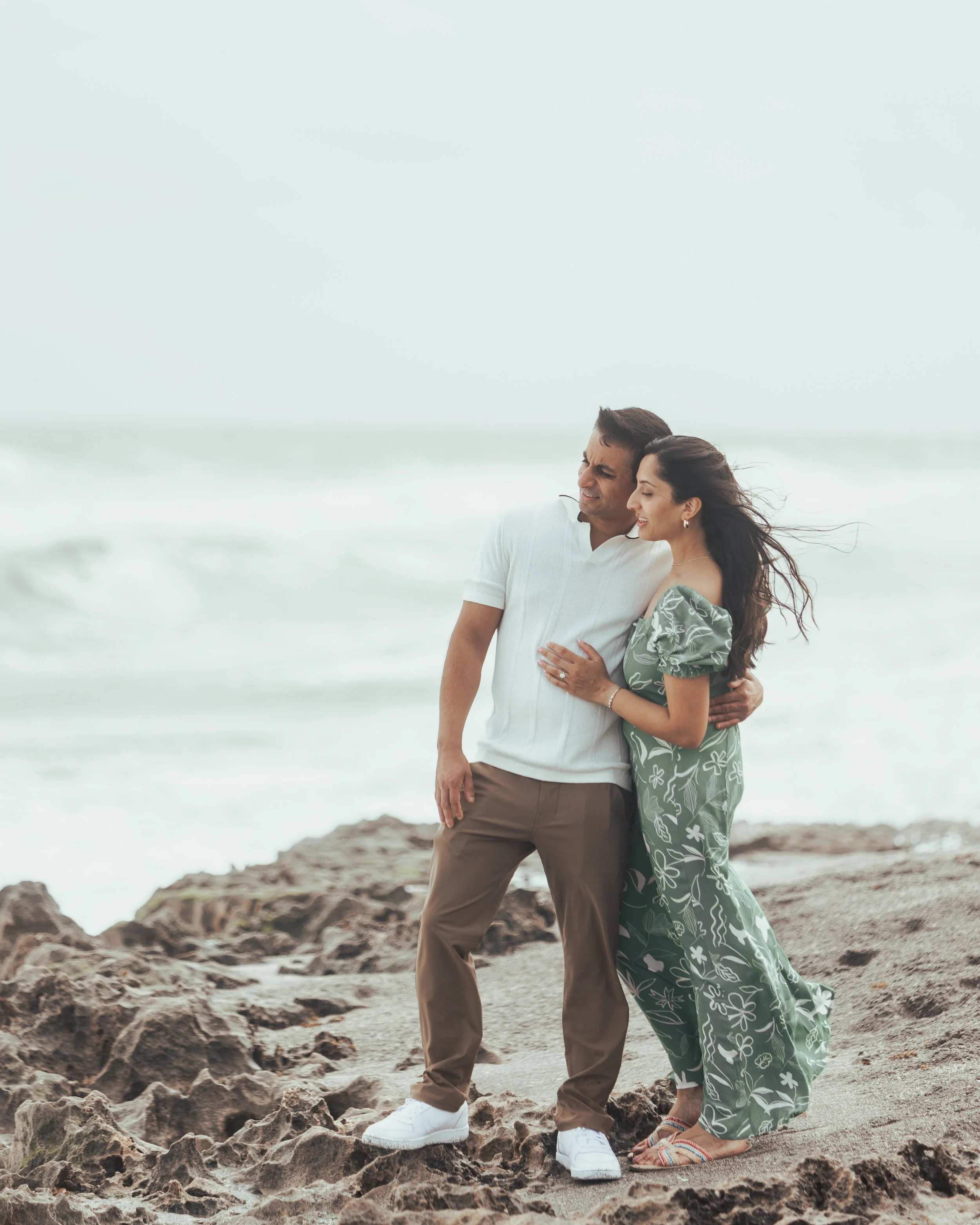 A couple standing on a rocky beach, holding each other, with the ocean in the background, smiling and enjoying the moment.