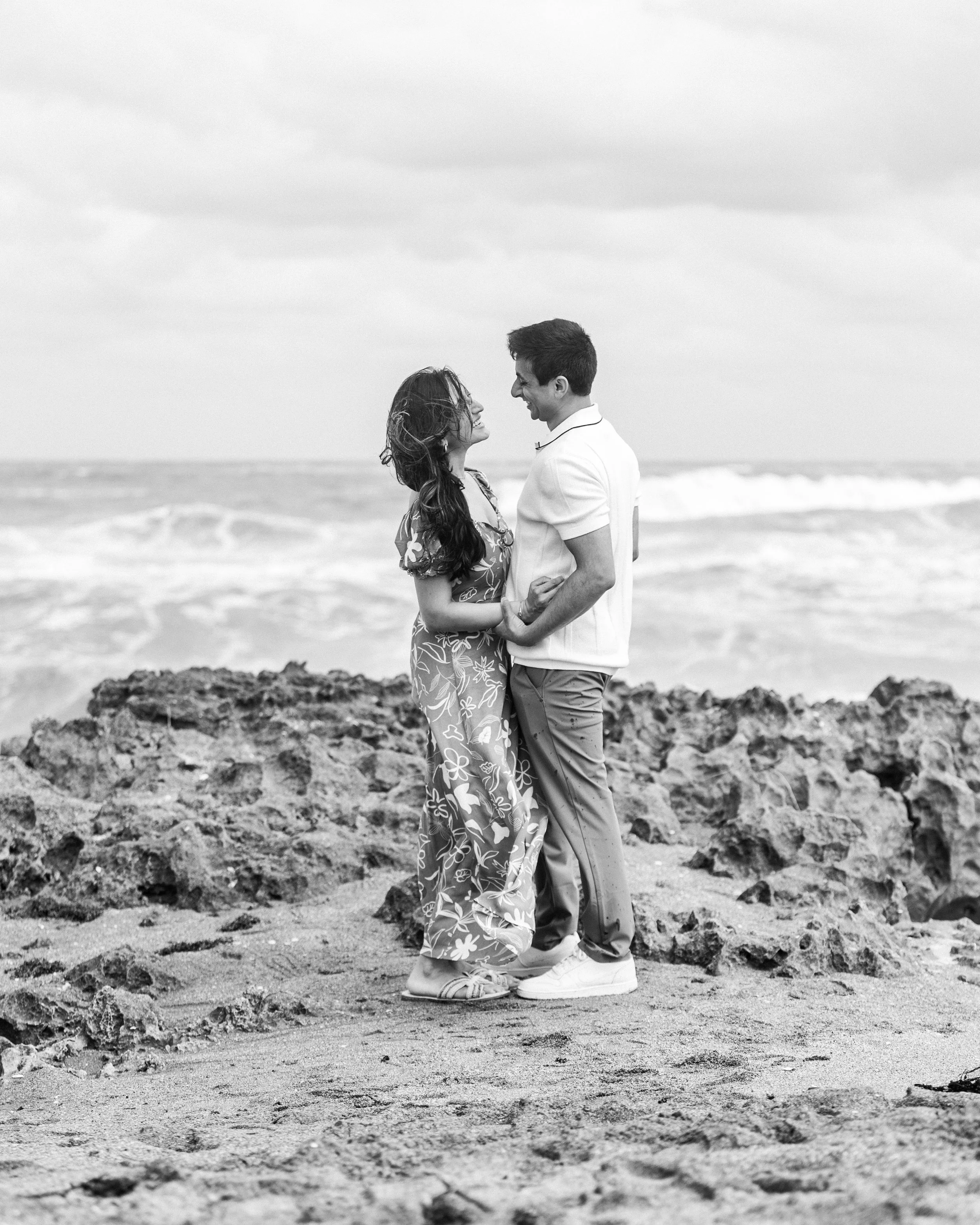 A black and white photo of a couple standing on a rocky beach, holding hands, and looking at each other with the ocean in the background.