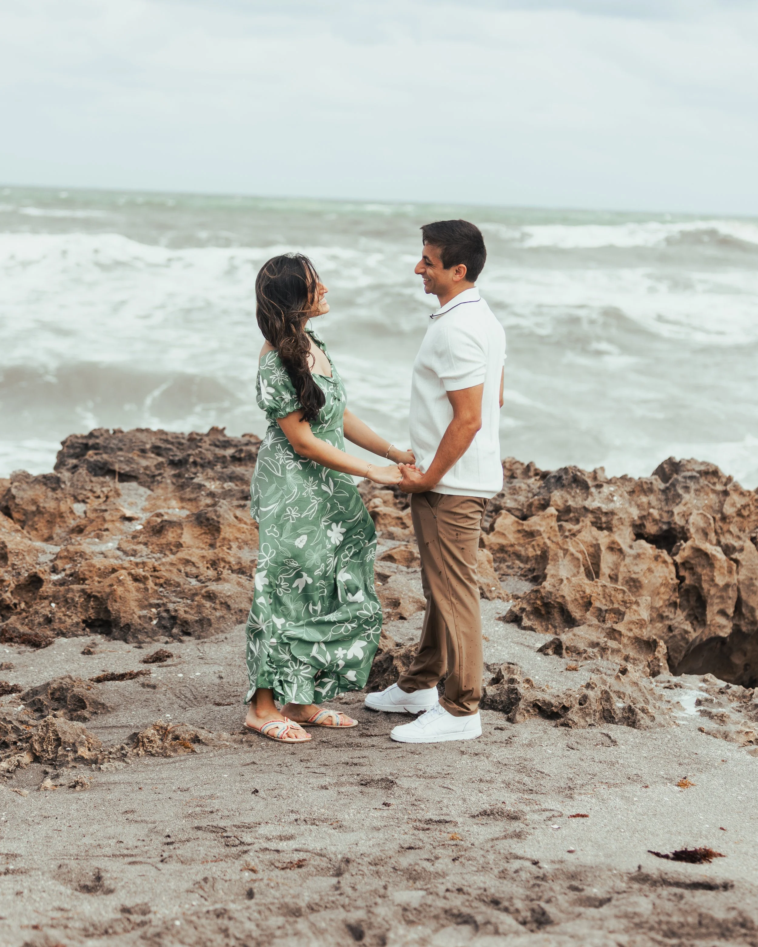A couple holding hands on a rocky beach, facing each other and smiling, with the ocean in the background.