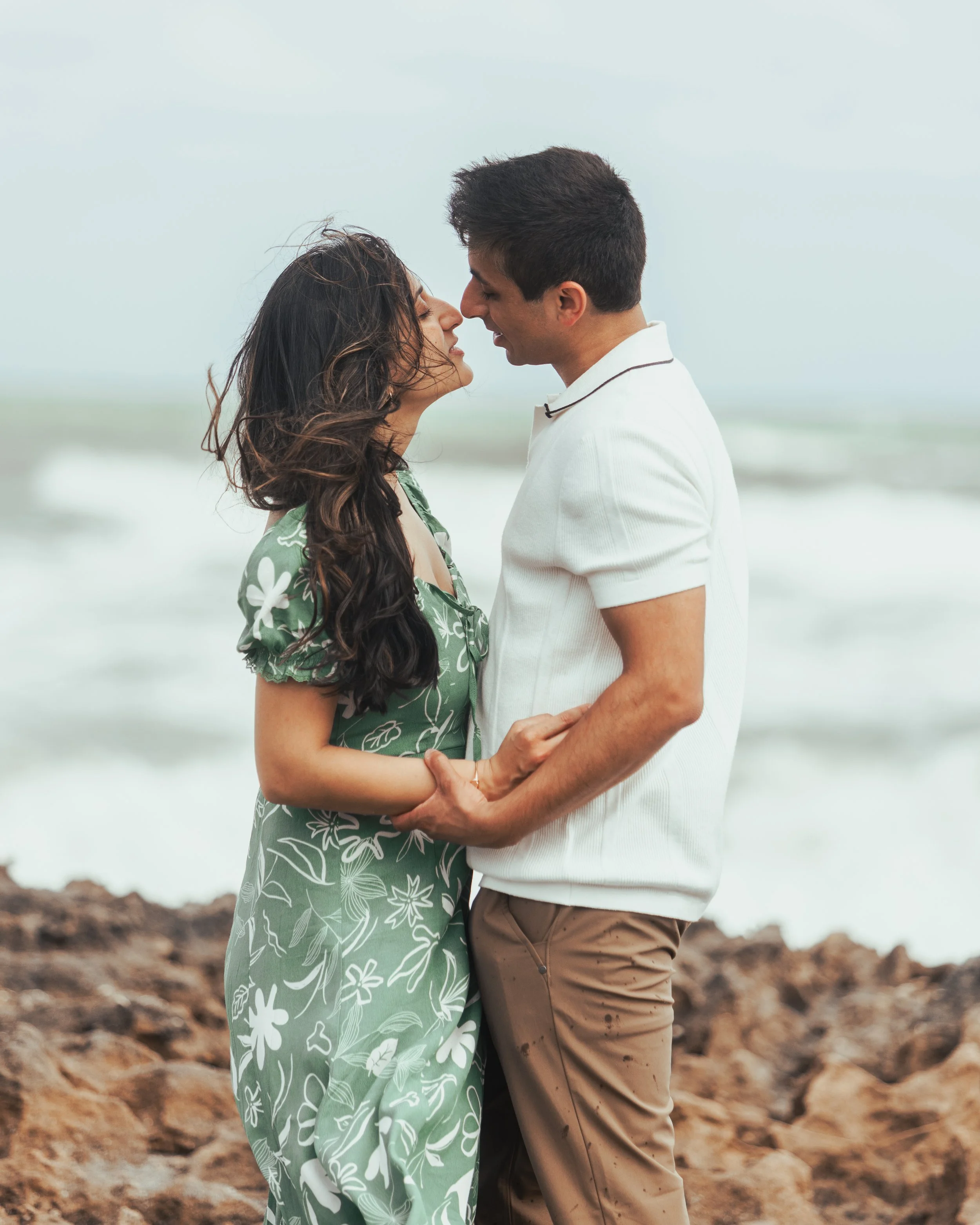 A couple stands close together on a rocky beach, facing each other, with the ocean in the background. They are holding hands and leaning in for a kiss.