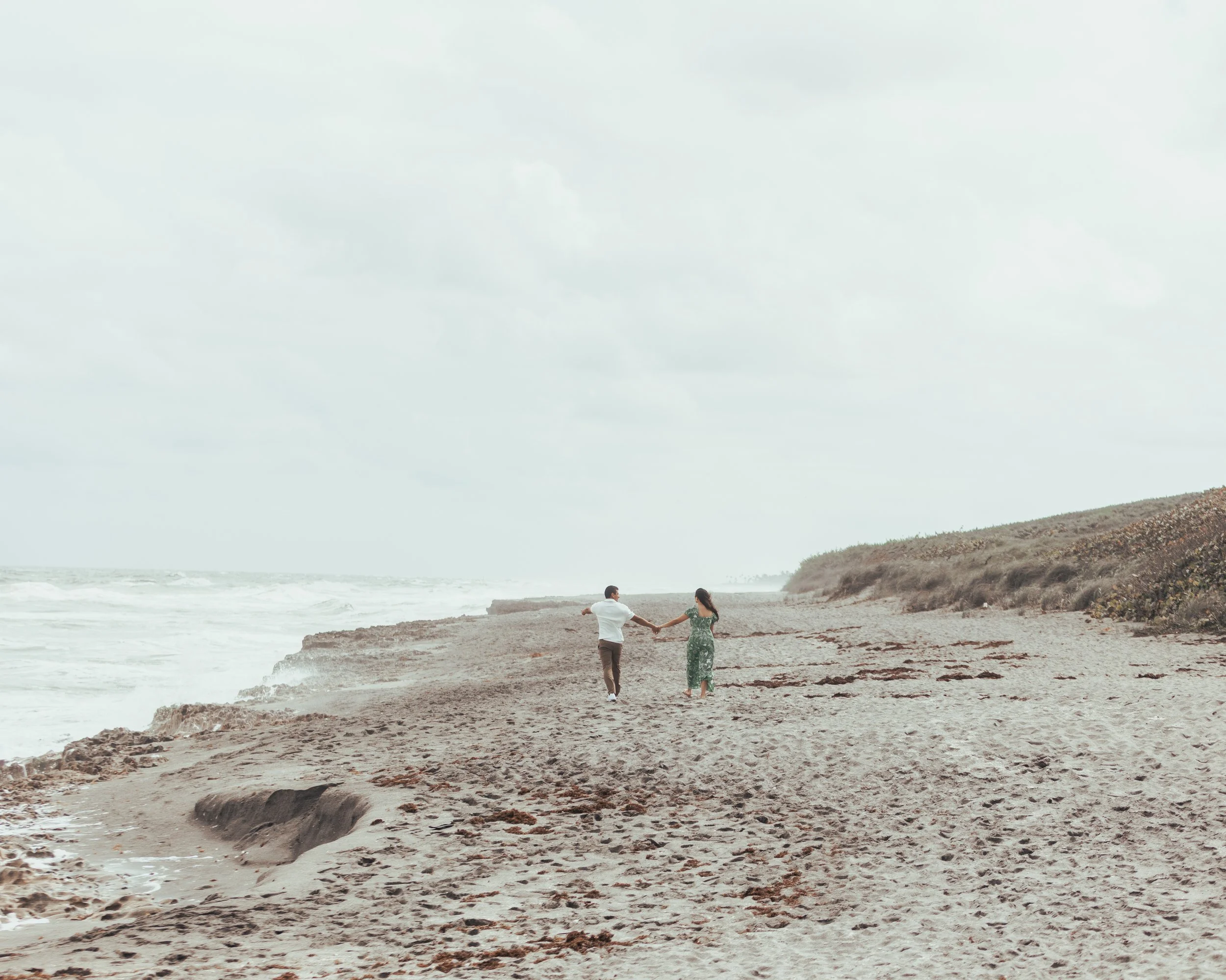 A couple holding hands walking on a sandy beach with waves and coast in the background.