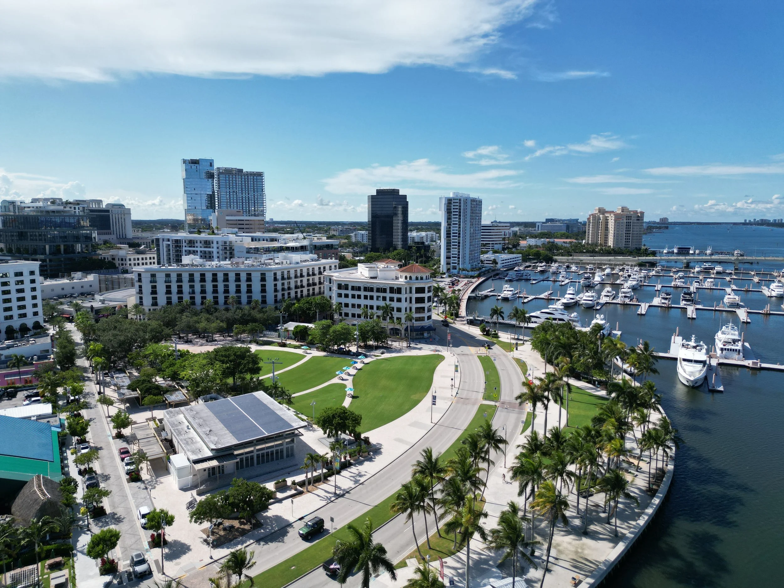 Aerial view of a city harbor with high-rise buildings, marina filled with yachts, and a park with walkways and palm trees under a partly cloudy sky.