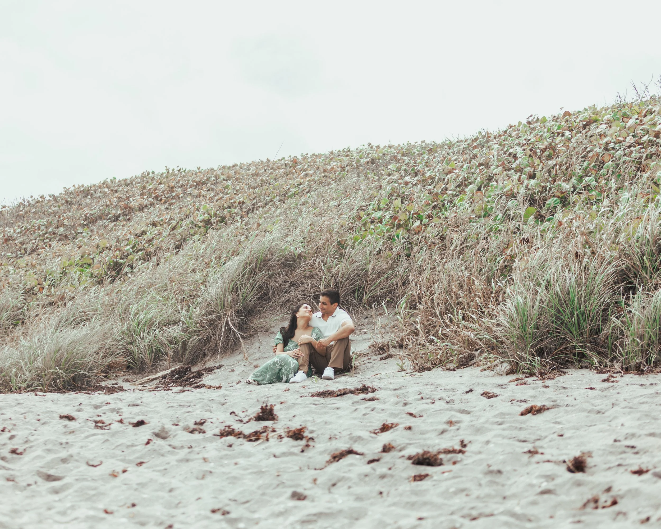 A couple sitting on the sand near grassy dunes at the beach.