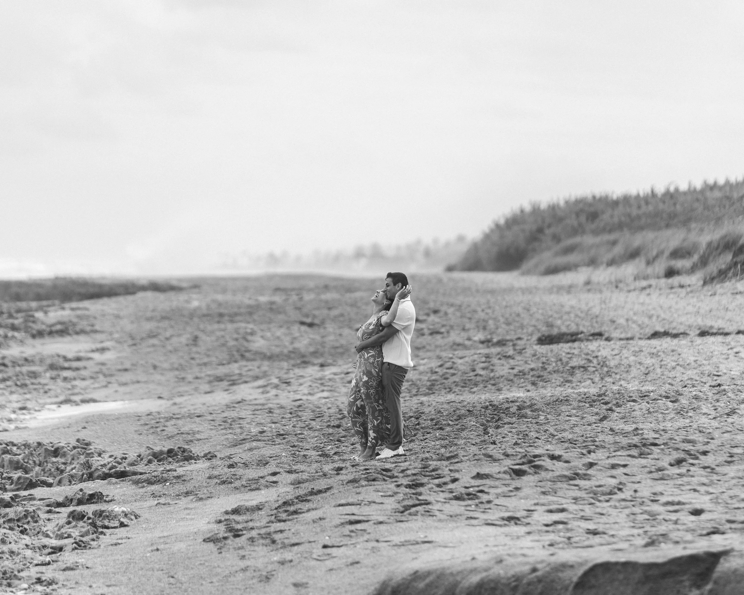 A couple hugging and smiling on a sandy beach with dunes and grass in the background, black and white photo.