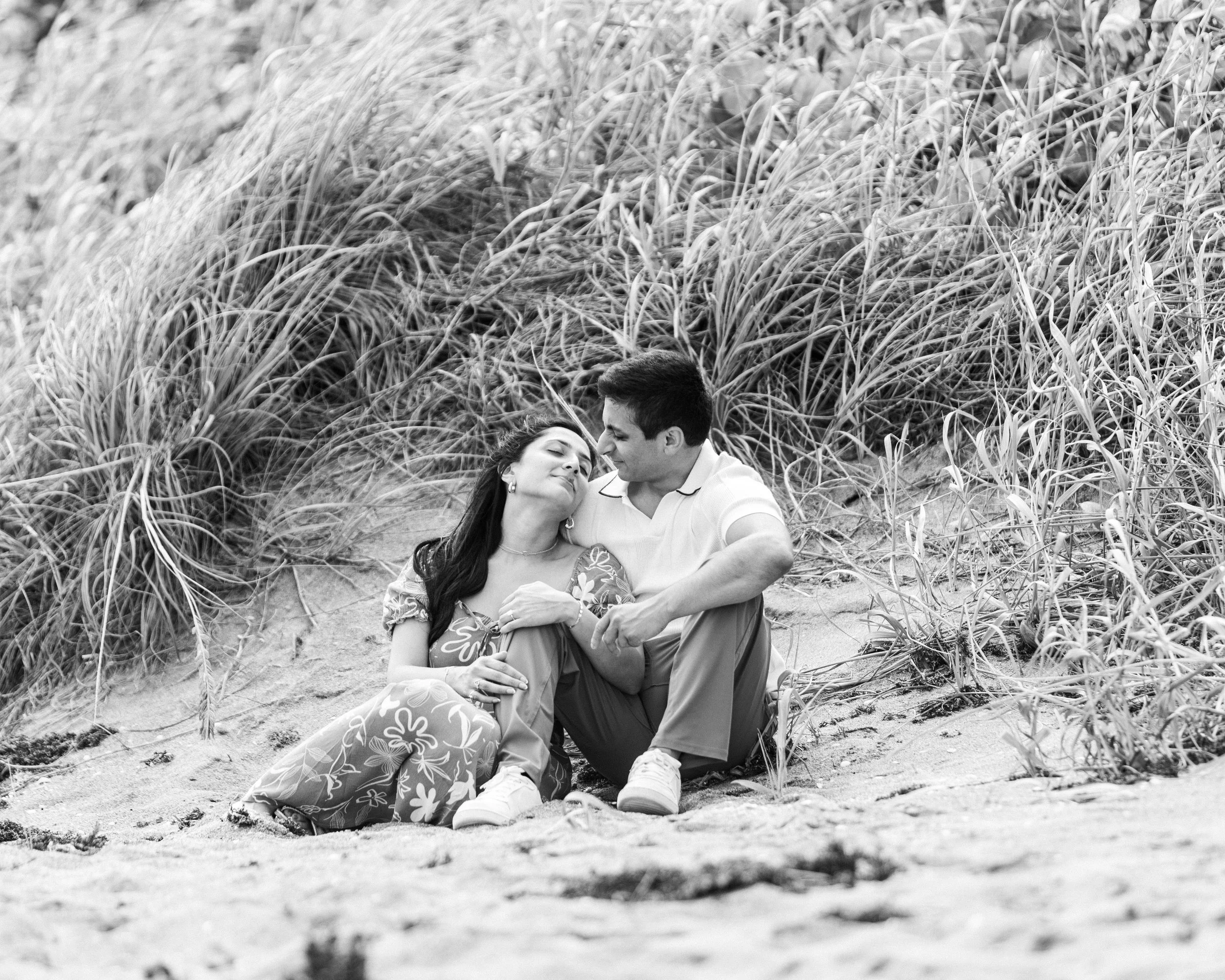 A black and white photo of a couple sitting on sand near tall grass, with the woman resting her head on the man's shoulder.