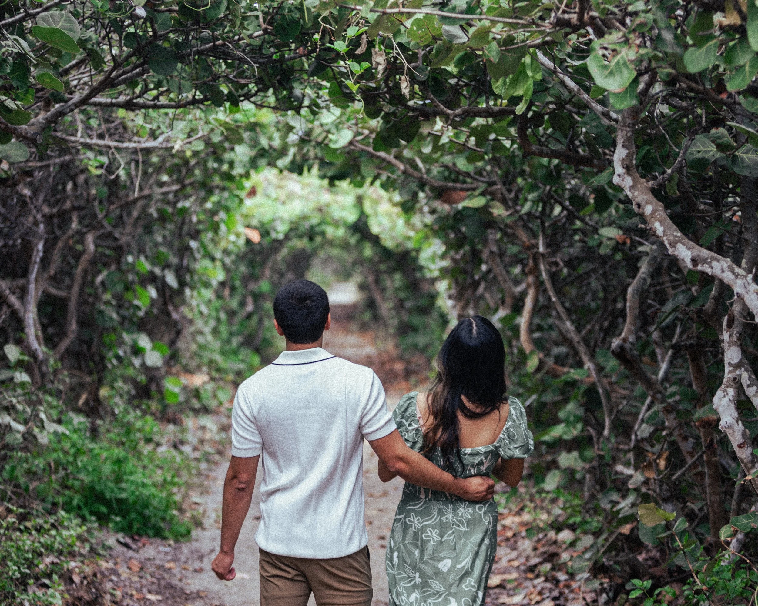 A man and woman walking closely together down a nature trail surrounded by dense green foliage, with the man's arm around the woman's shoulder.