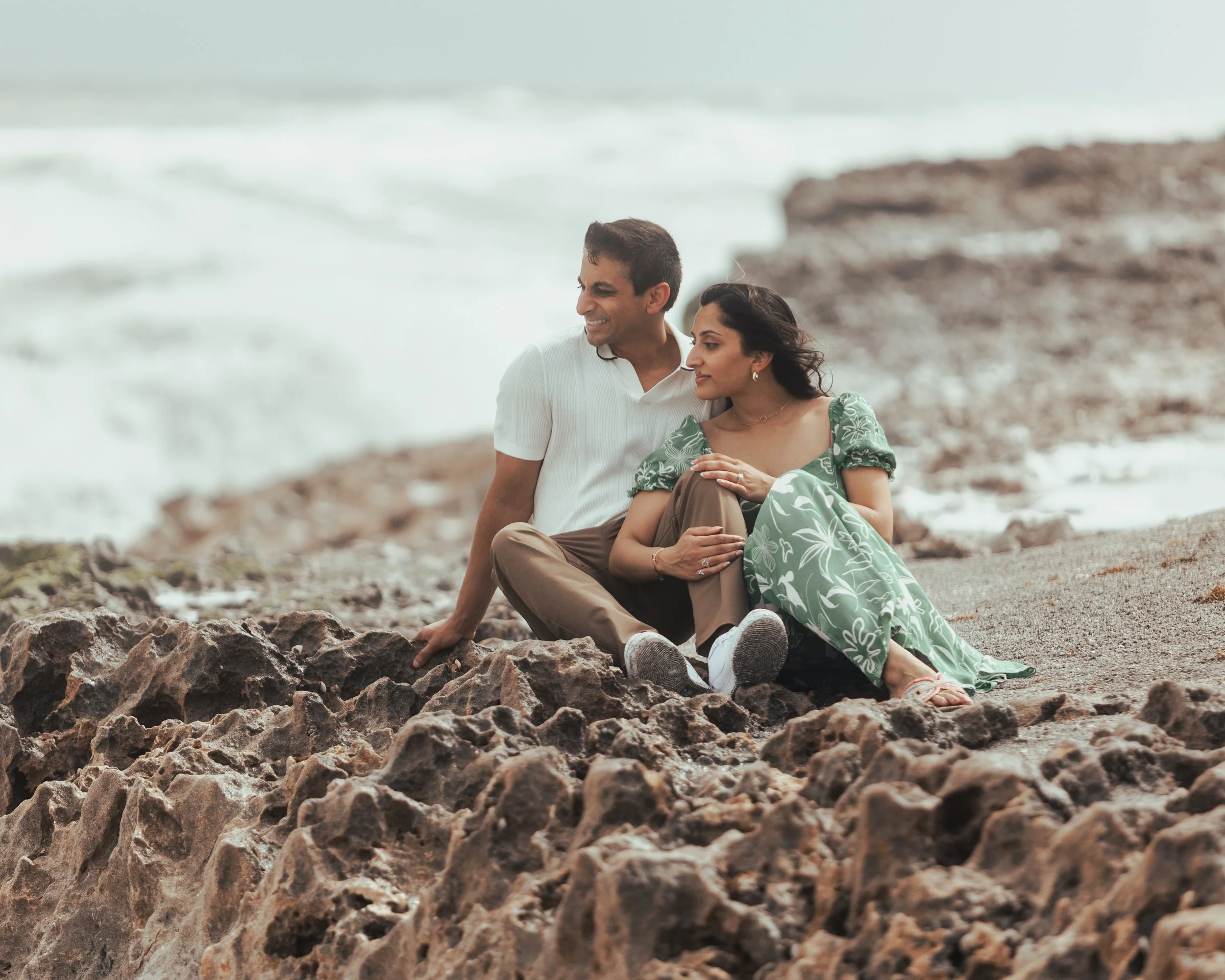 A couple sitting on rocks by the beach, with the ocean in the background, smiling and relaxing.