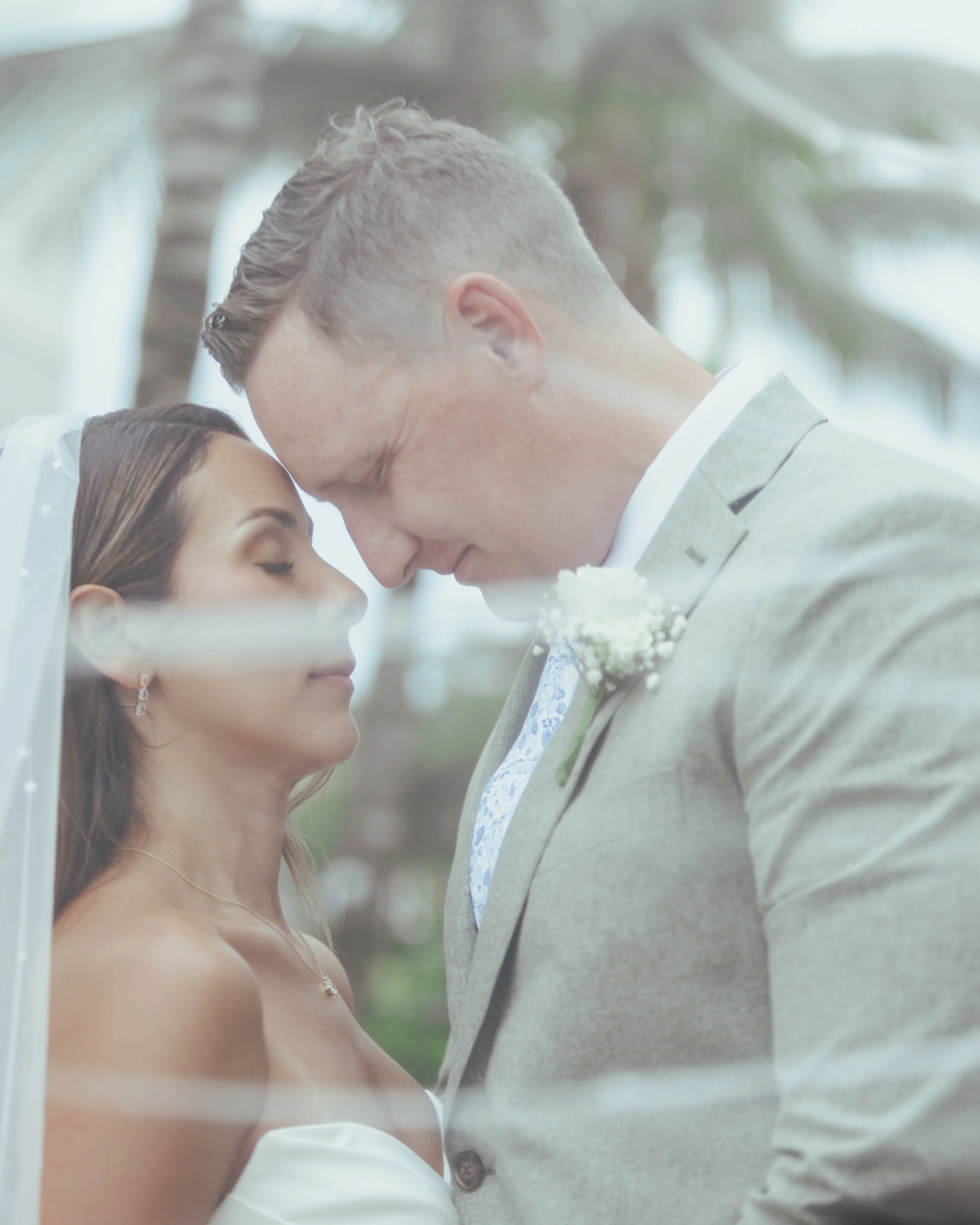 A bride and groom with their foreheads touching, eyes closed, during their wedding ceremony outdoors with palm trees in the background.