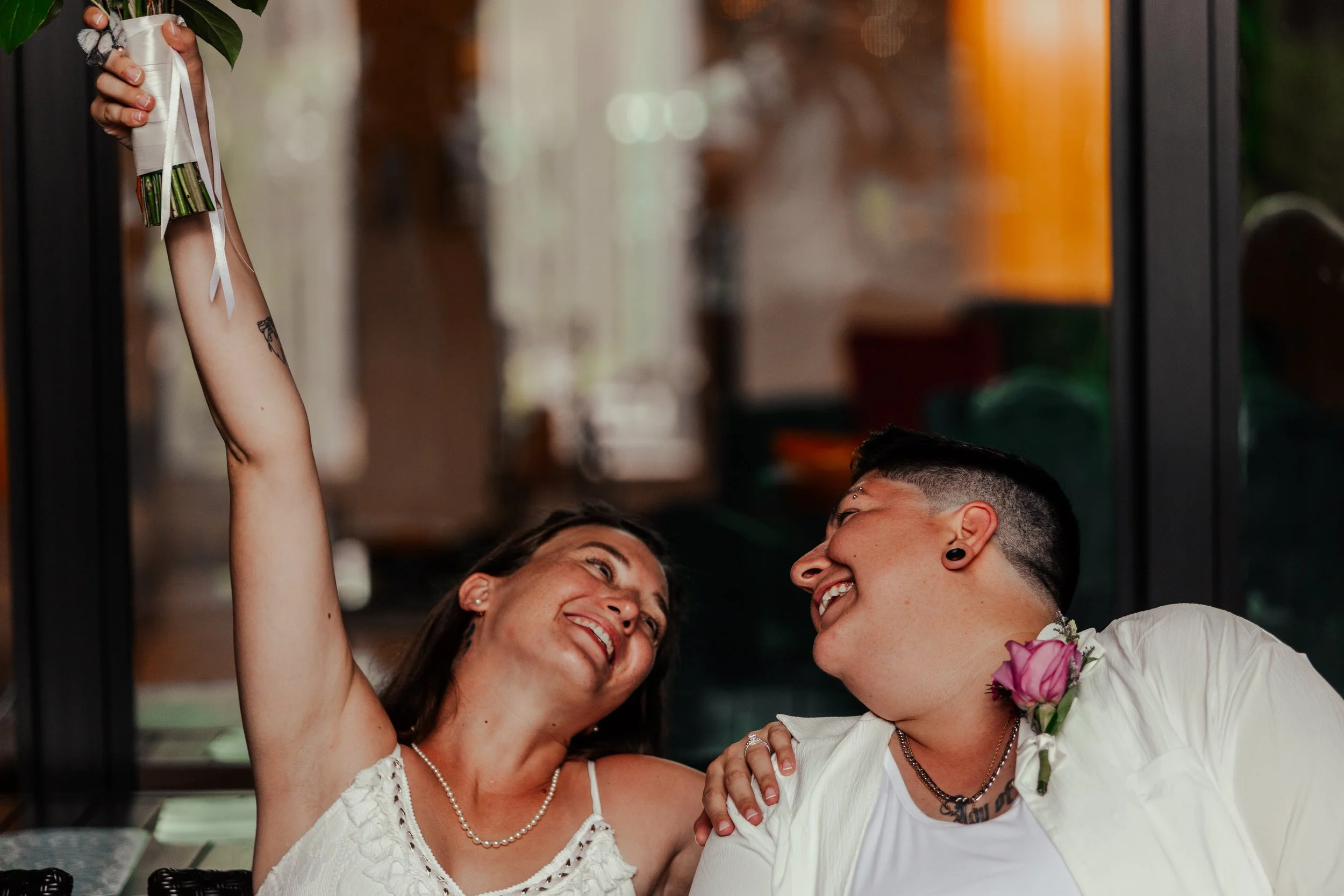 Two people smiling and looking at each other, one holding a bouquet of flowers, inside a room with large glass windows.