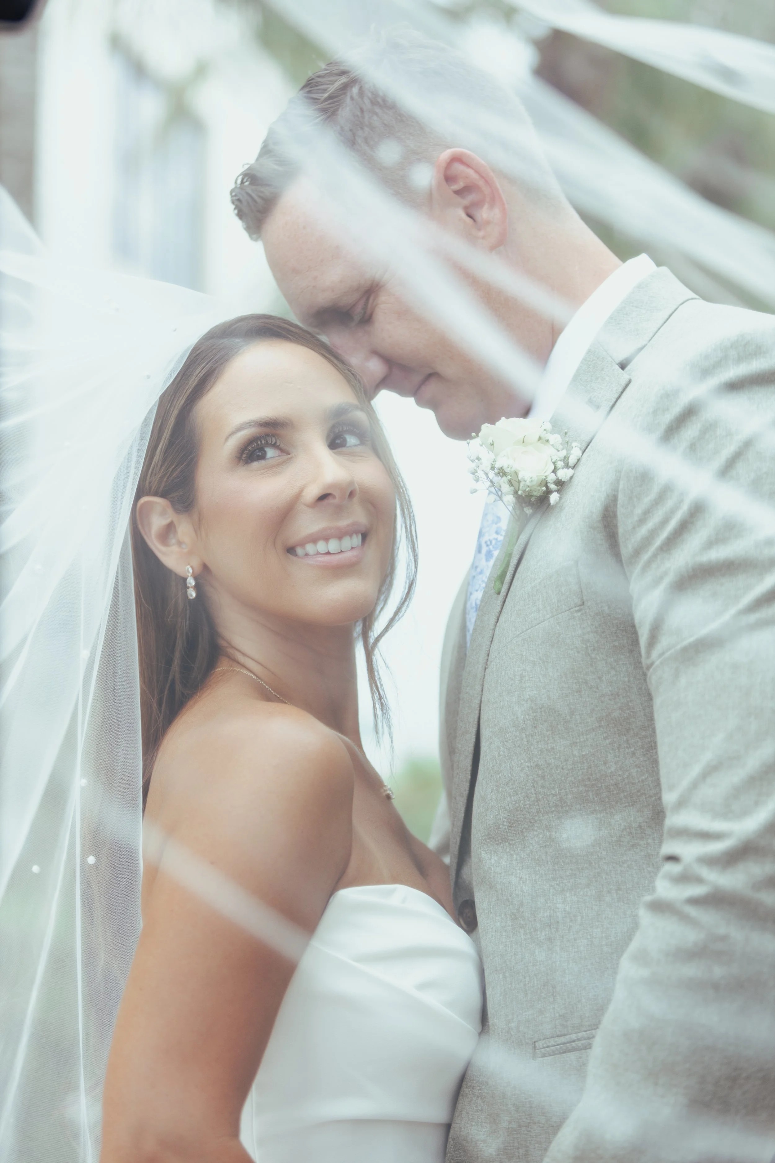 A bride and groom close-up at their wedding, with the bride smiling and the groom gently resting his forehead on hers.